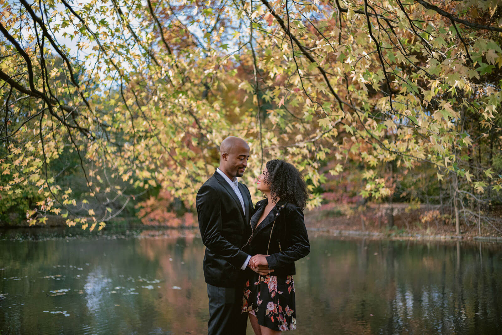 The engaged couple is standing close together in front of a lake in Brooklyn. NYC Image by Jenny Fu Studio  