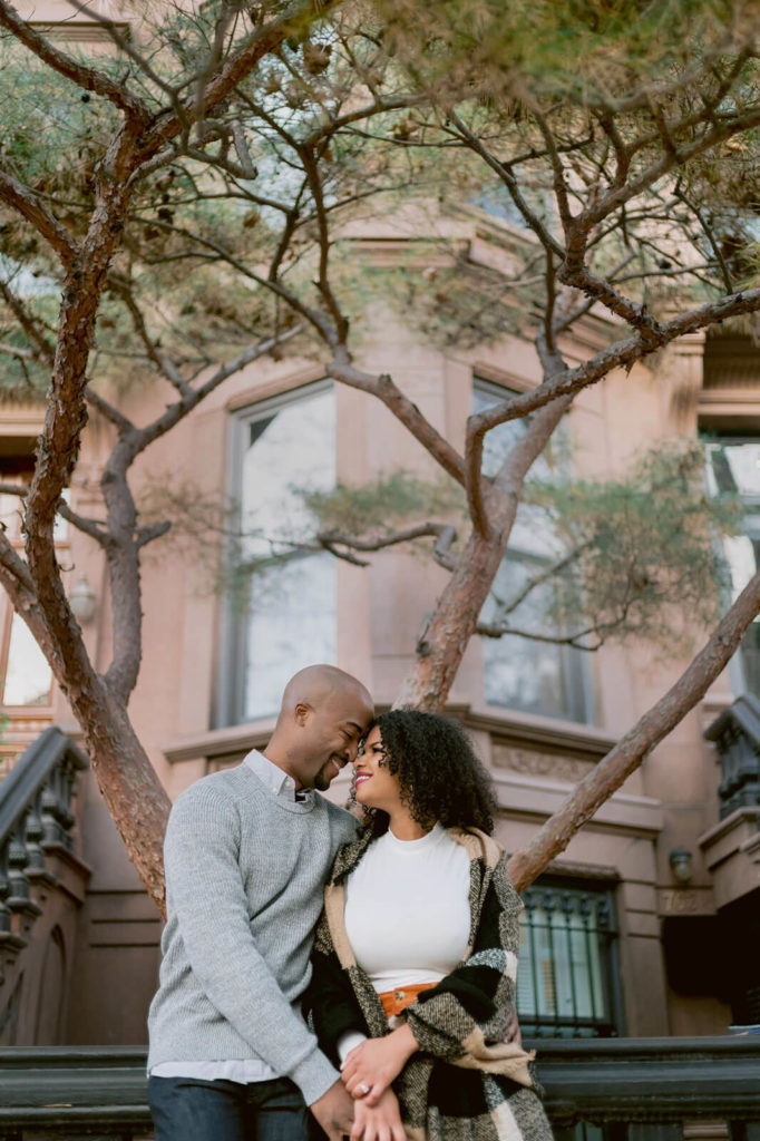 The engaged couple is romantically staring at each other in front of a house in Brooklyn. NYC Christmas Engagement Photos by Jenny Fu Studio