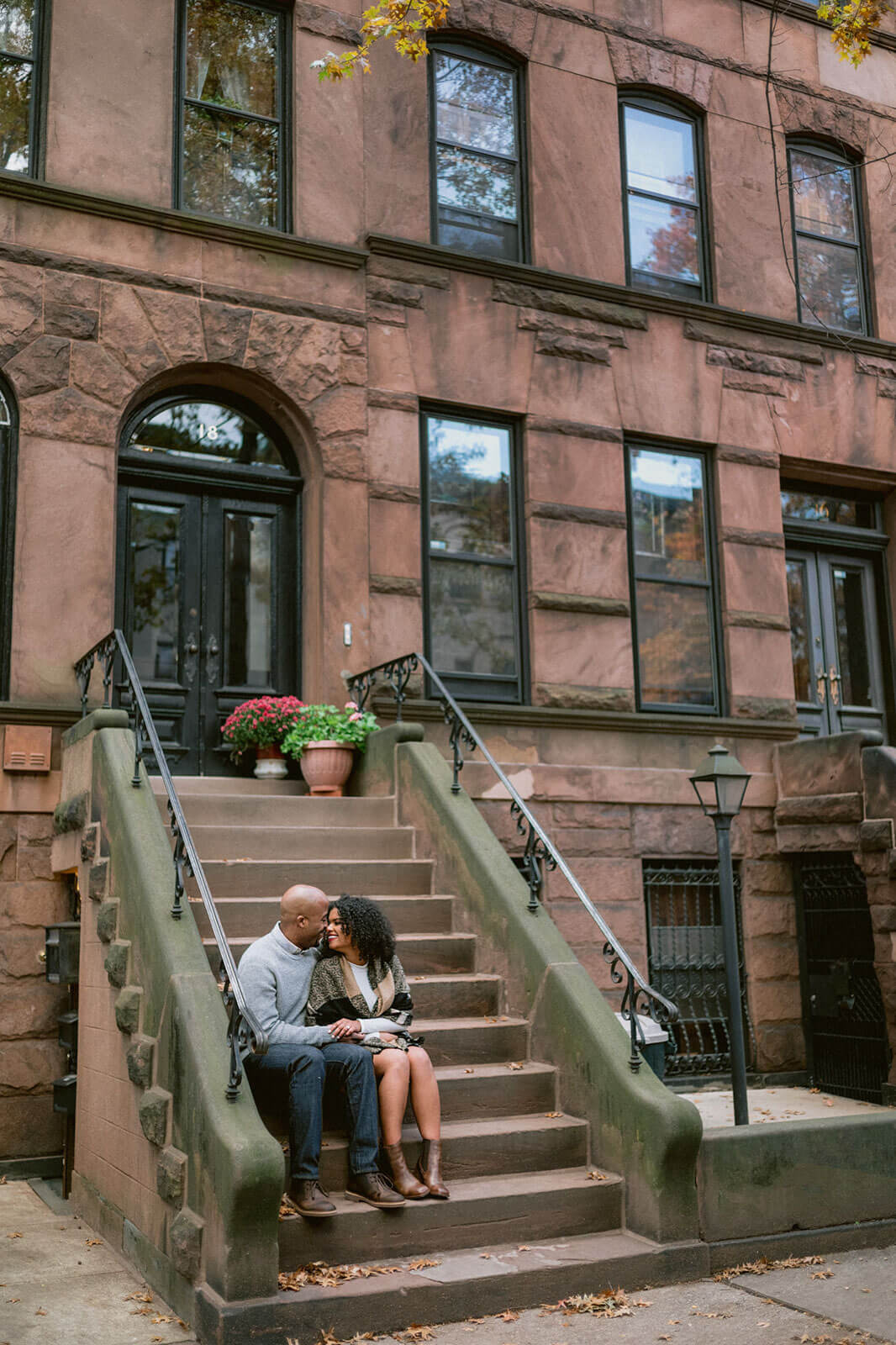 The engaged couple is sitting in a staircase on the streets of Brooklyn. NYC Christmas Engagement Photos by Jenny Fu Studio