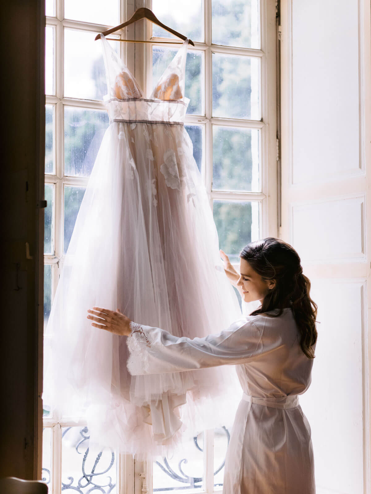 The bride, wearing a robe, is happily touching her wedding dress. Image by Destination Wedding Photographer Jenny Fu