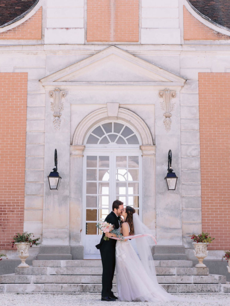 The bride and groom kiss in front of a chateau in France. Image by Destination Wedding Photographer Jenny Fu