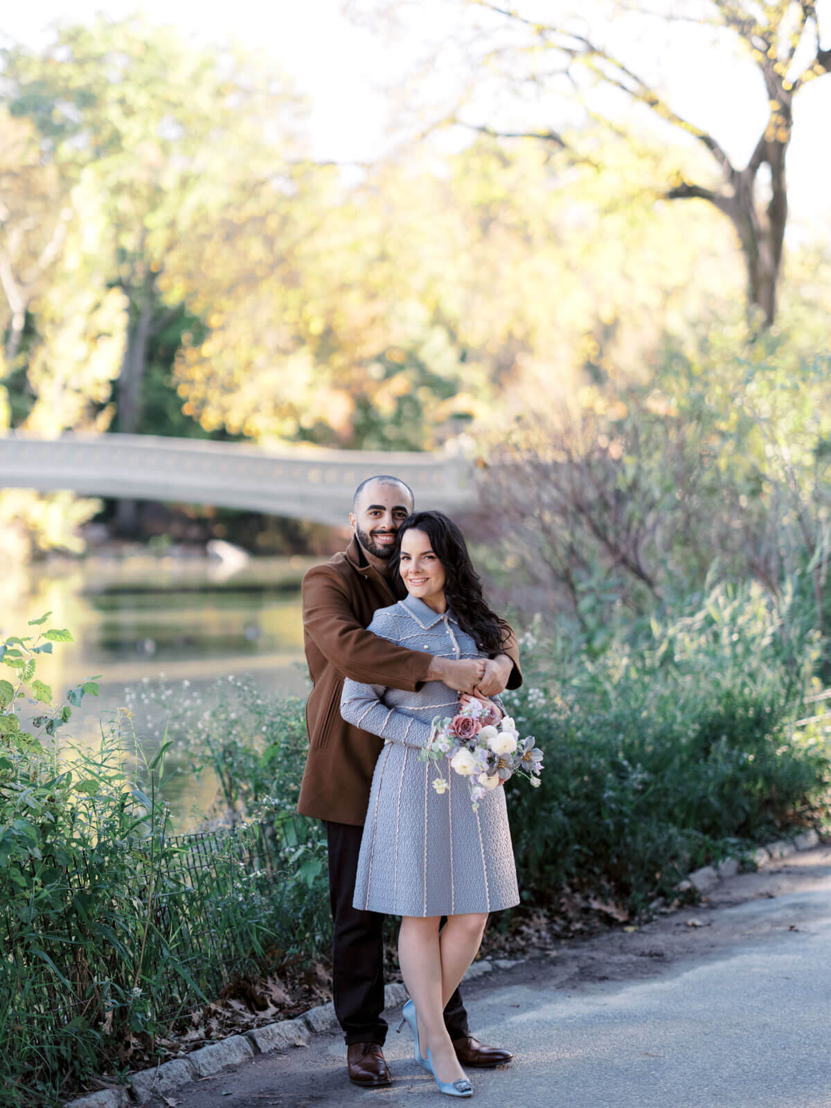 The engaged man is lovingly hugging his fiancée from the behind. At the background is a bridge and lots of trees.  Engagement image by Jenny Fu Studio