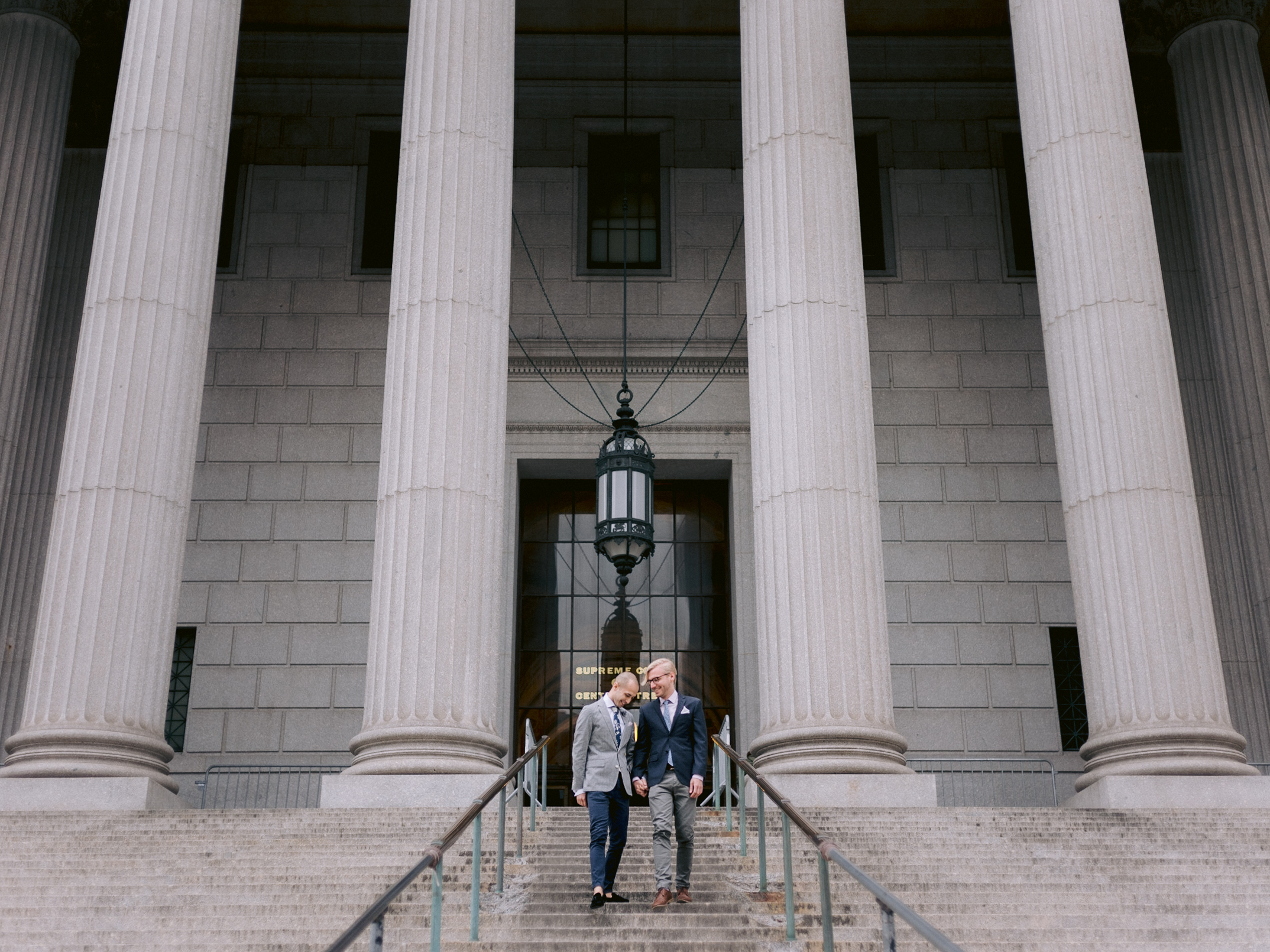 The two grooms are standing on the grand staircase entrance of the city hall. NYC City Hall elopement image by Jenny Fu Studio 
