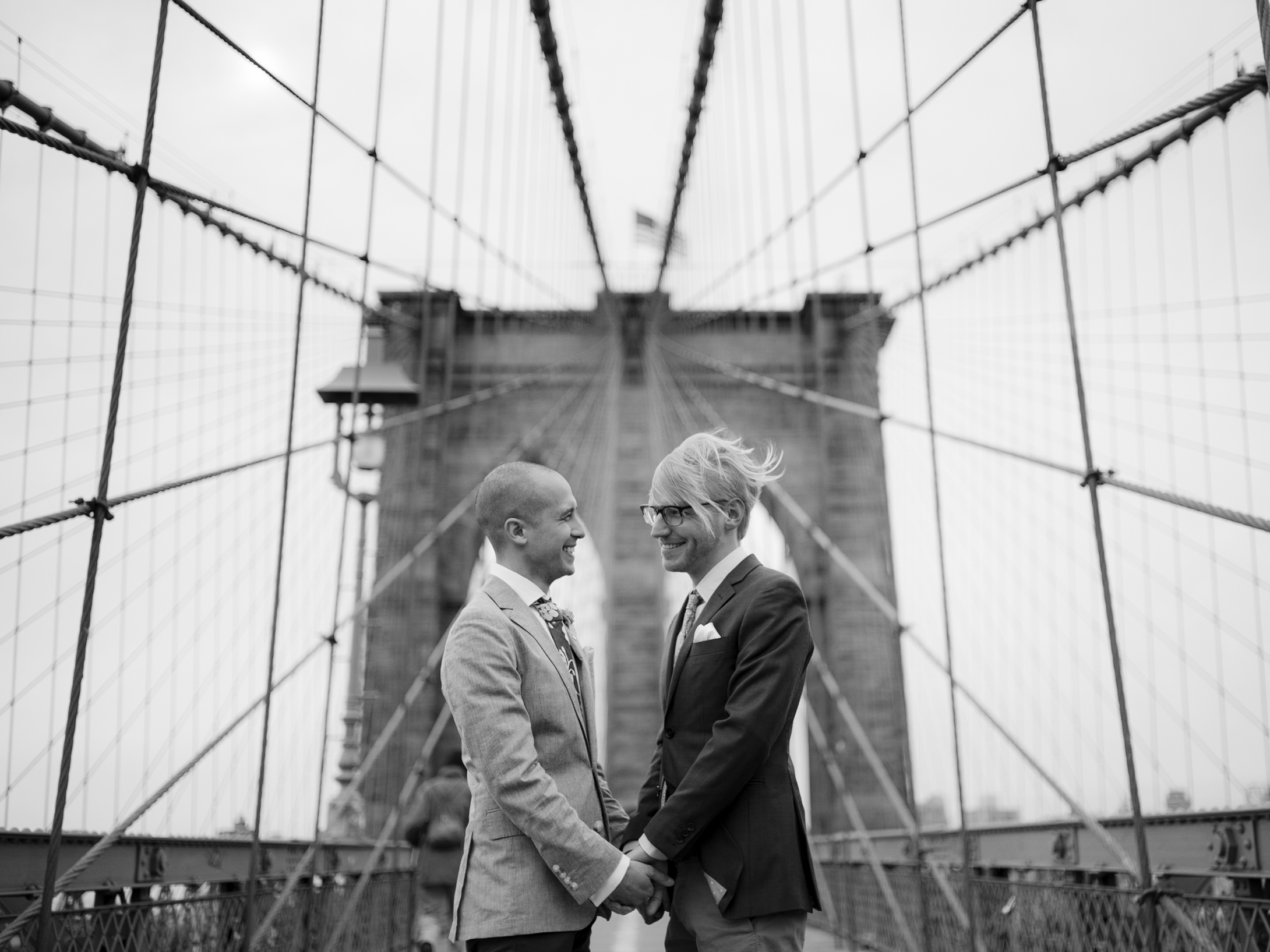 The two grooms are looking at each other happily while holding hands at Brooklyn Bridge. Elopement image by Jenny Fu Studio