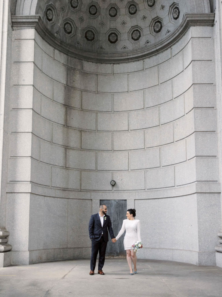 The bride and groom are in front of a curved wall in NYC City Hall.