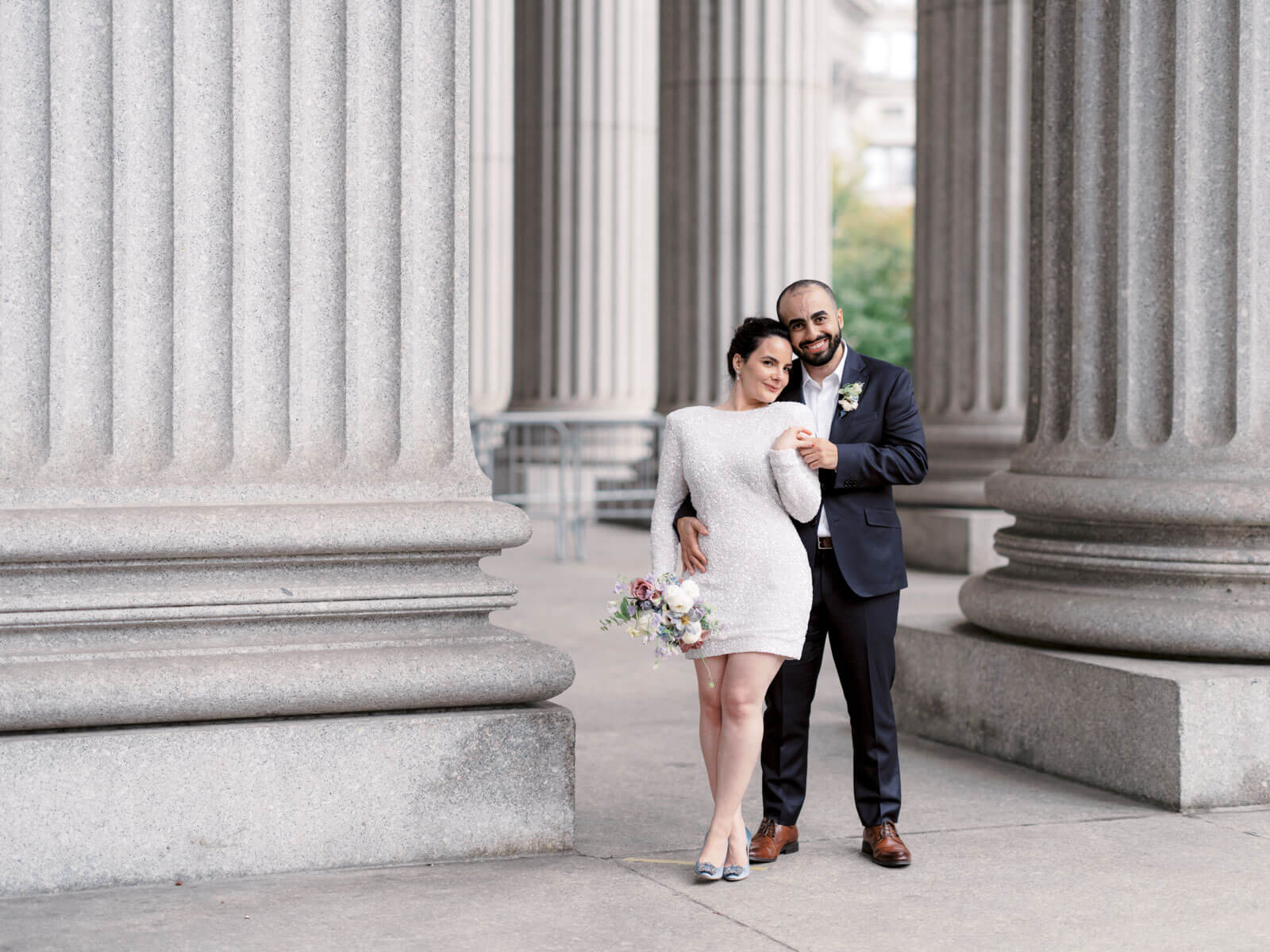 The bride and groom are standing close to each other amidst the large columns at NYC City Hall. NY city hall elopement image by Jenny Fu Studio
