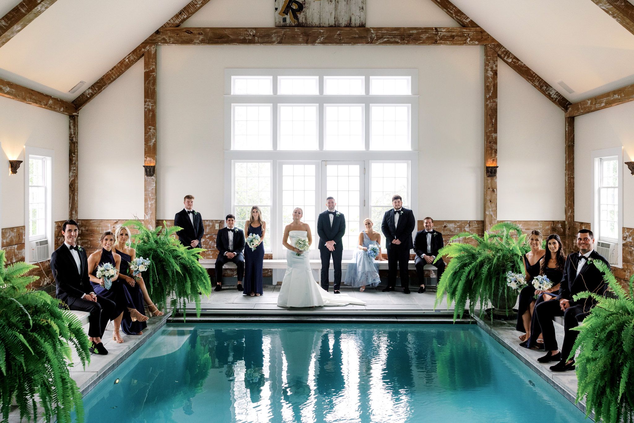 The bride, groom, groomsmen, and bridesmaids pose in a large hall with a swimming pool. Image by destination wedding photographer Jenny Fu
