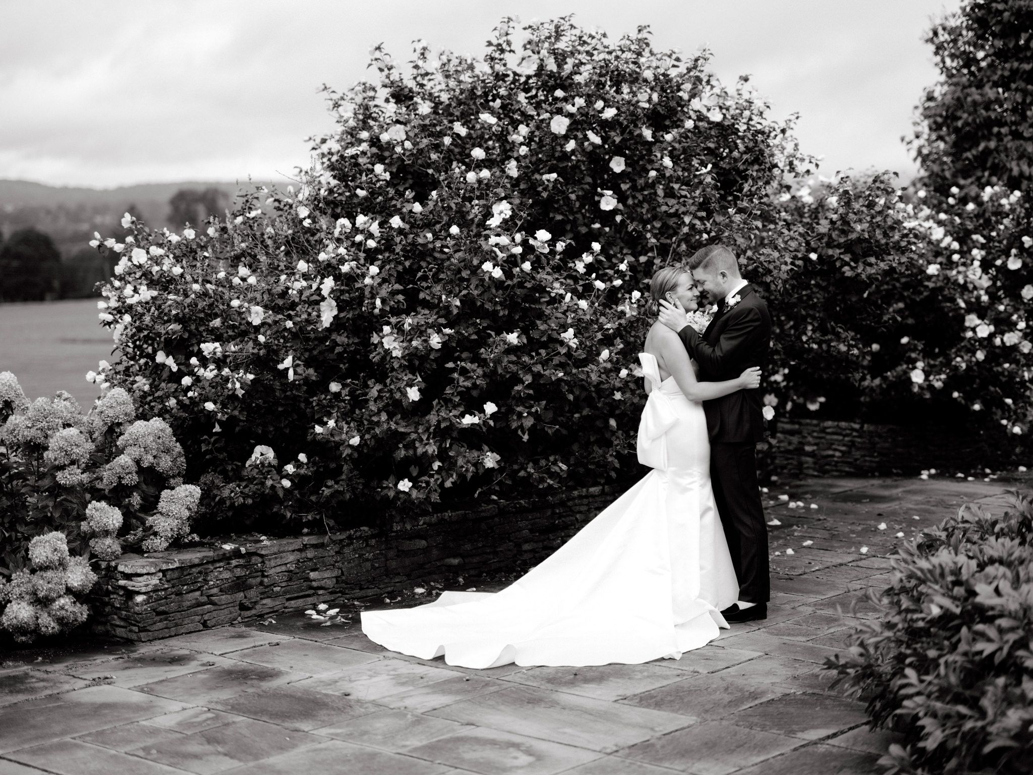 Black and White photo of the bride and groom standing romantically close together amidst many flowers. Destination image by Jenny Fu Studio
