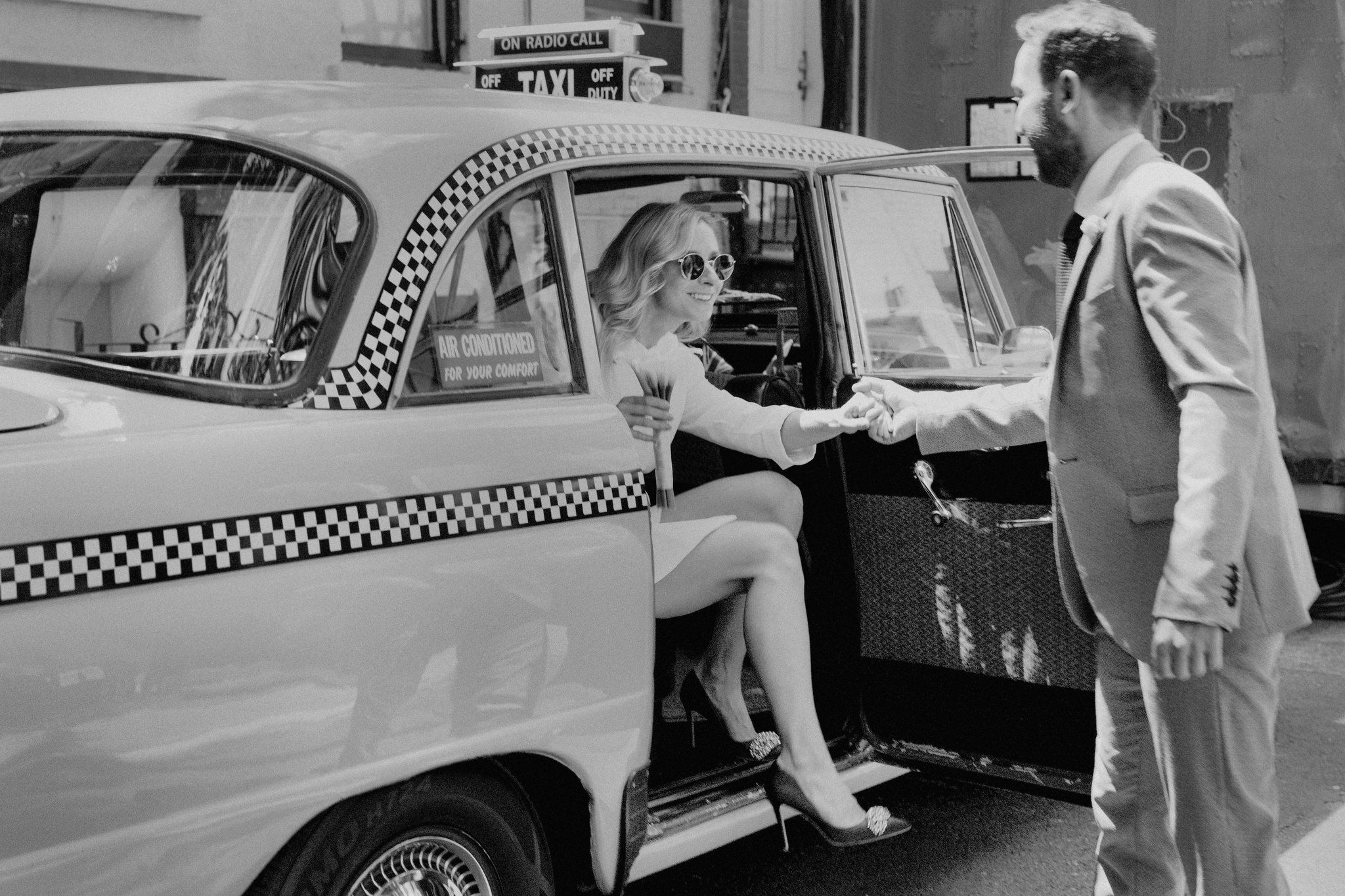 Black and white photo of the bride going out of a classic NYC yellow cab as the groom holds her hands. Image by Jenny Fu Studio