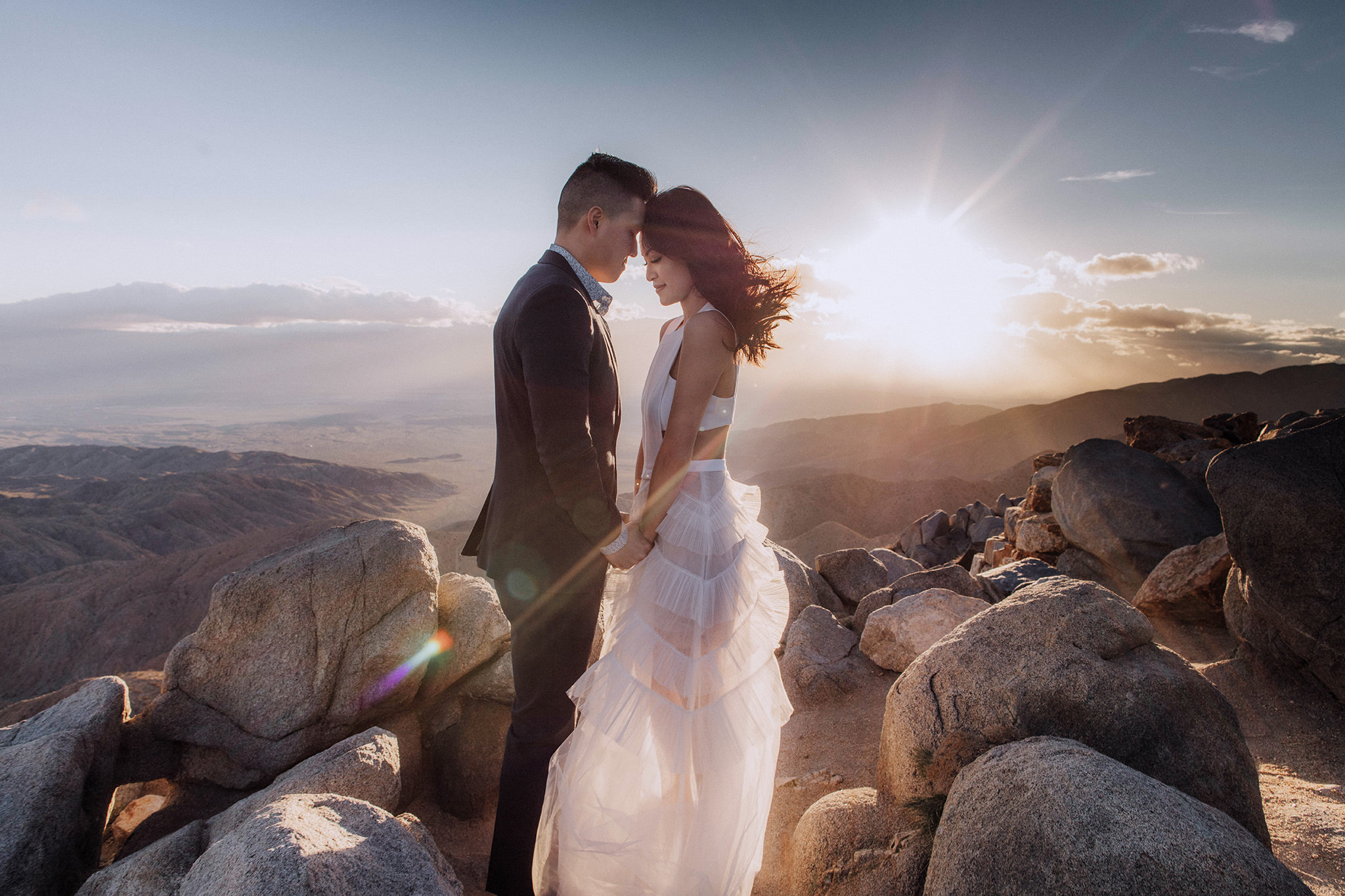 The engaged couple is standing, heads touching and holding hands in the middle of big rocks, with mountains and sunset in the background. Image by Jenny Fu Studio