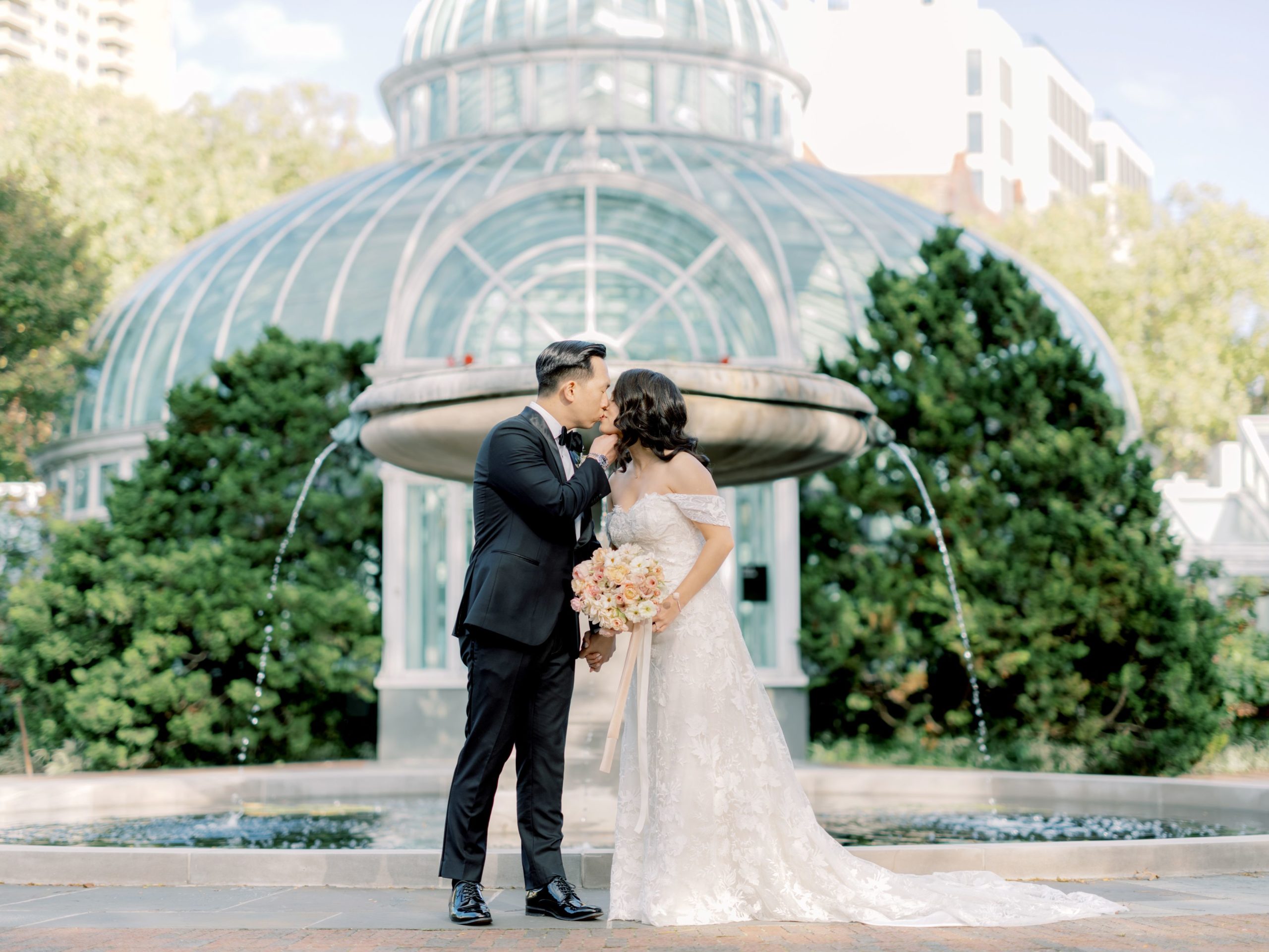 The bride and the groom are kissing each other in front of a beautiful fountain. Hiring a wedding planner image by Jenny Fu Studio