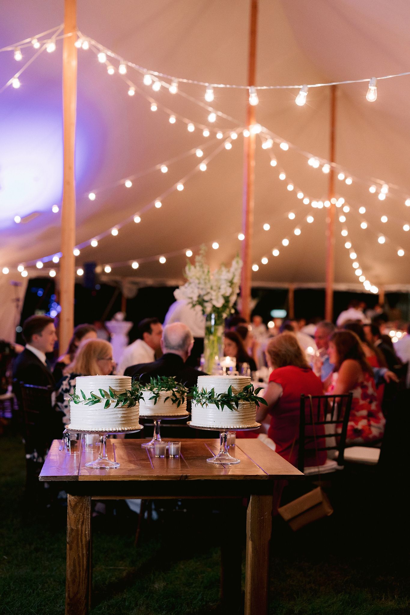 Wedding cake on a table with the guests eating in the pretty background. Wianno Club, Cape Cod, Osterville, MA. Beach wedding venue image by Jenny Fu Studio