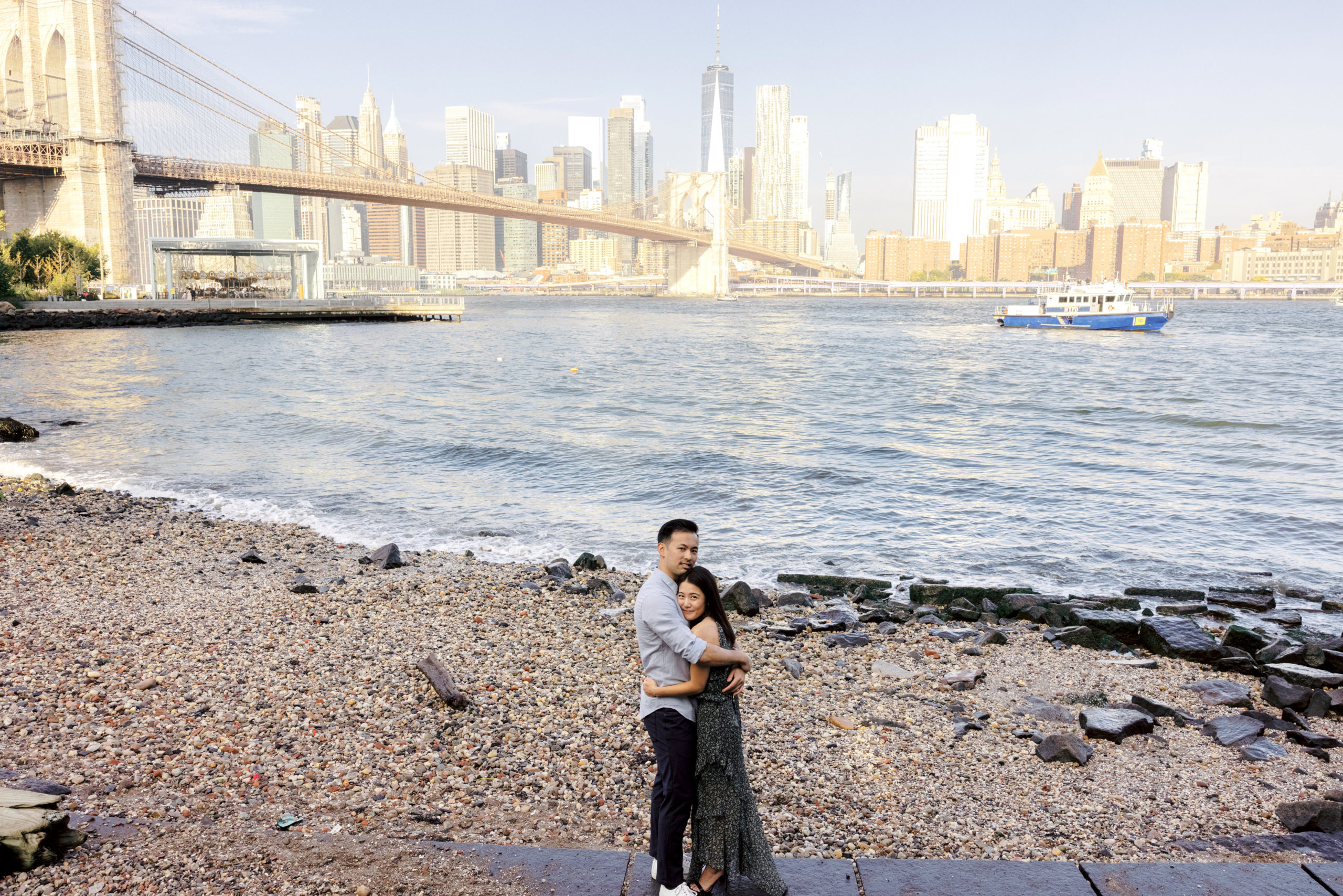 The engaged couple are hugging each other with the East River and Brooklyn Bridge in the background. Editorial engagement image by Jenny Fu Studio