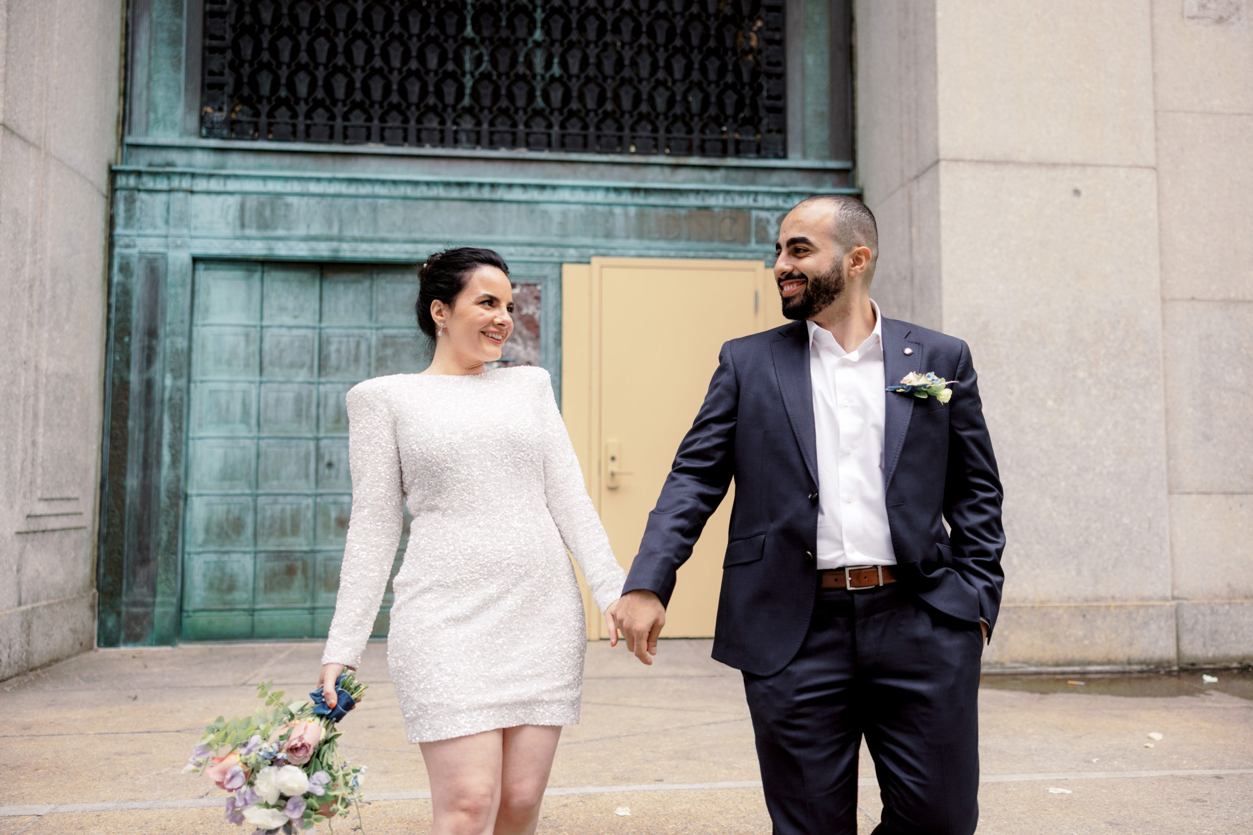The bride and groom are smiling at each other with the city hall in the background. Editorial wedding image by Jenny Fu Studio NYC