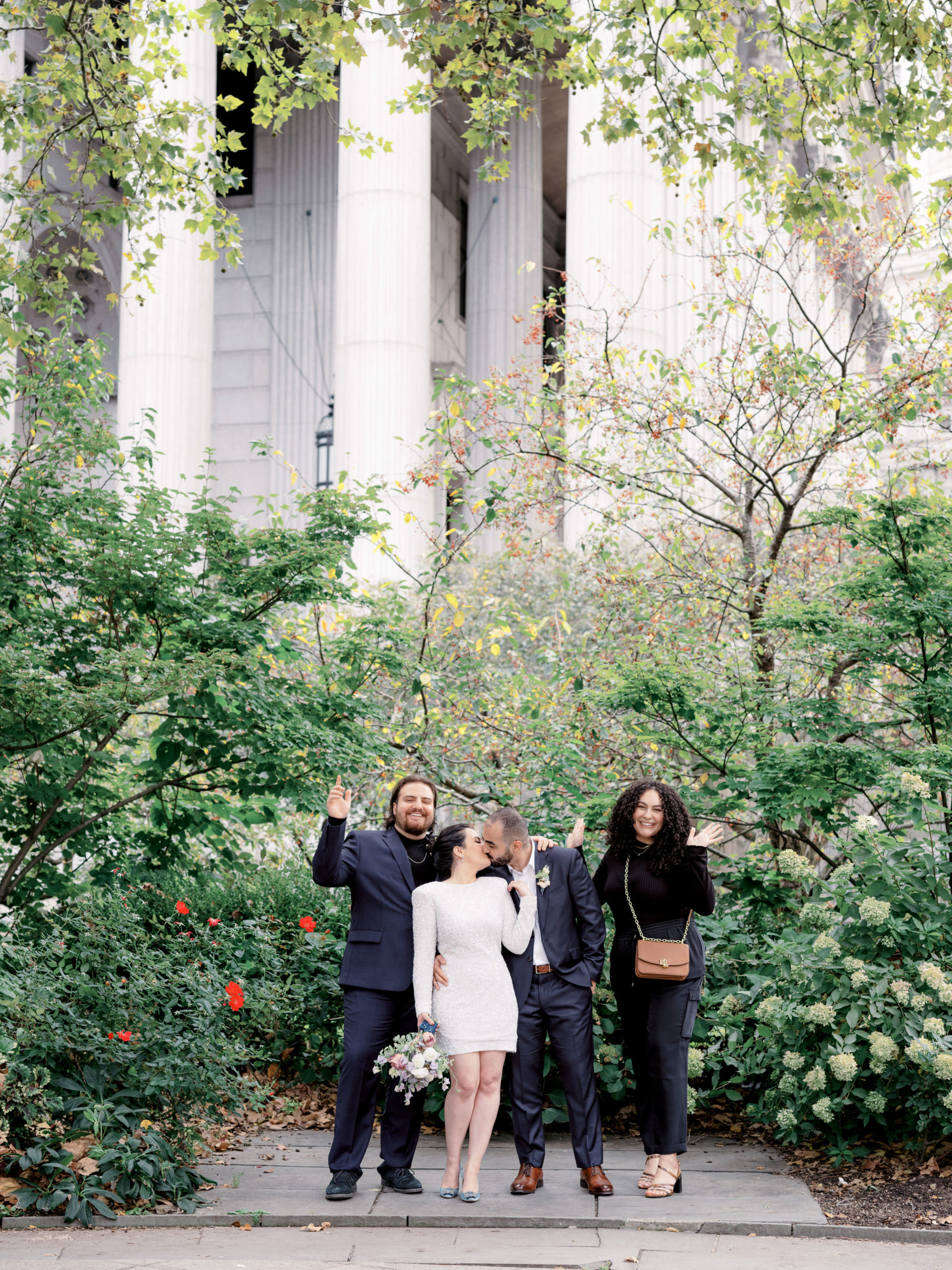 The bride and groom are kissing, their friends, smiling, with trees in the background. Image by Jenny Fu Studio NYC