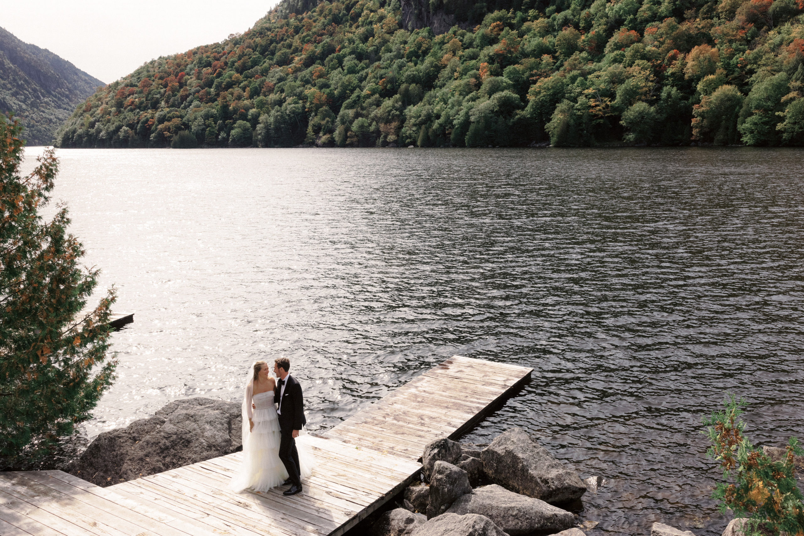 The bride and the groom are standing on a dock beside a lake at The Ausable Club, New York. Destination Wedding Image by Jenny Fu Studio