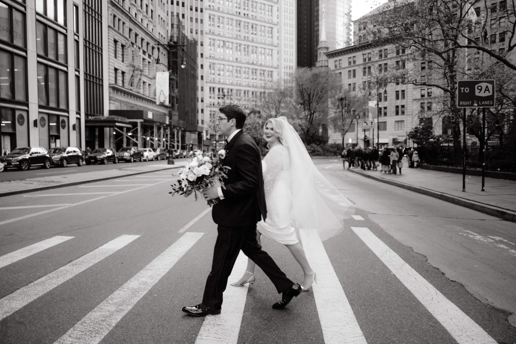 Black and white editorial photo of the bride and groom crossing the street with NYC city view background. Image by Jenny Fu Studio