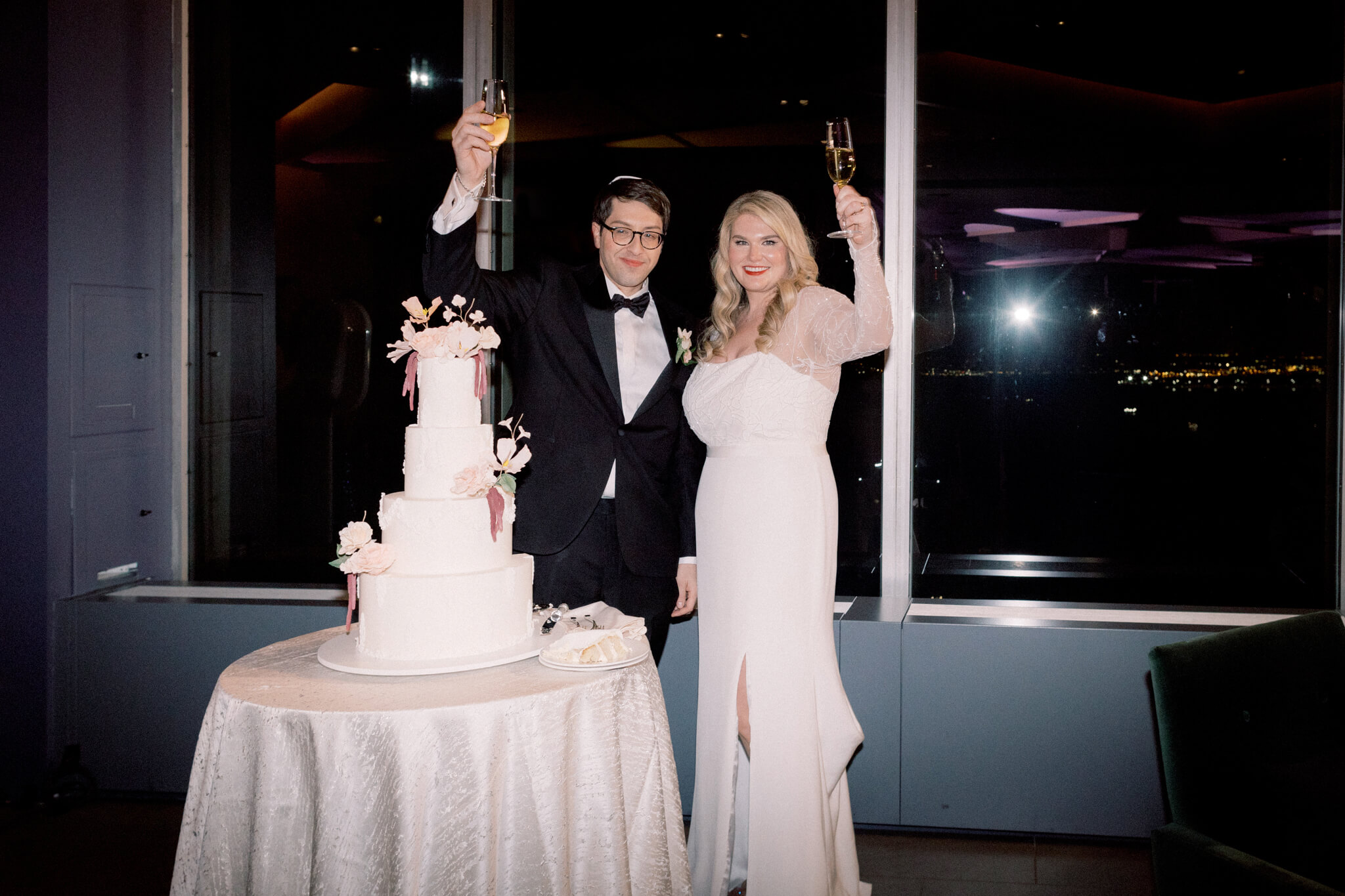 The bride and groom are holding their champagne glasses up in the air, with the wedding cake in front of them at Manhatta Restaurant, New York. Image by Jenny Fu Studio