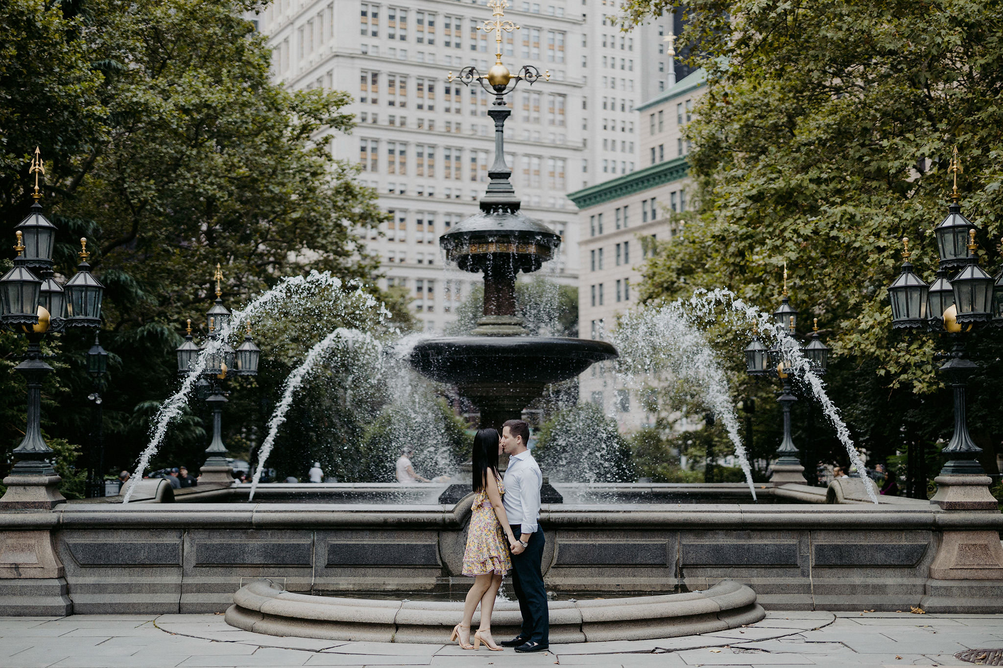 The engaged couple is kissing in front of a fountain at Central Park. Editorial NYC engagement image by Jenny Fu Studio