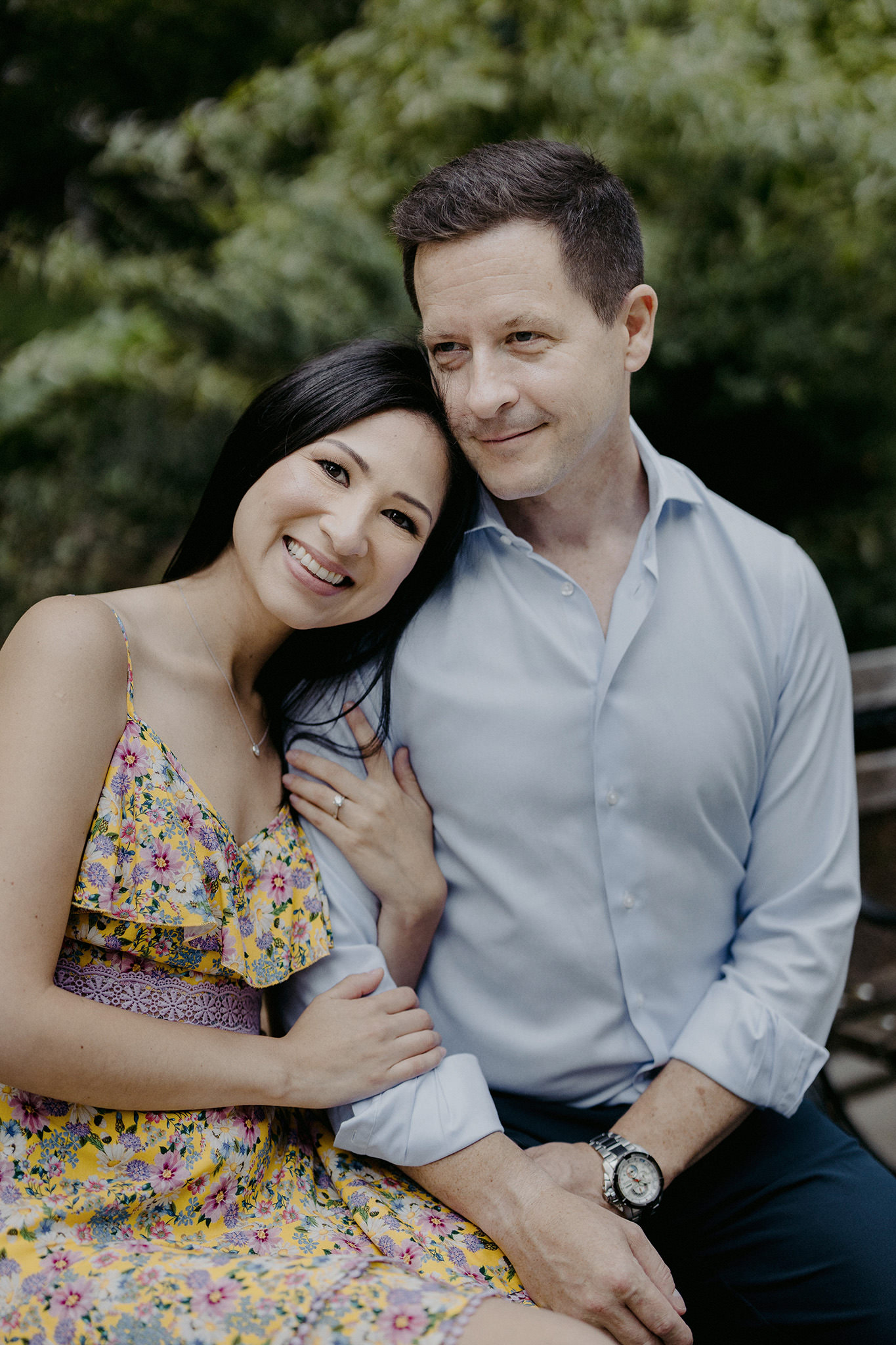 The engaged couple is smiling while sitting on a bench at Central Park. NYC summer engagement image by Jenny Fu Studio
