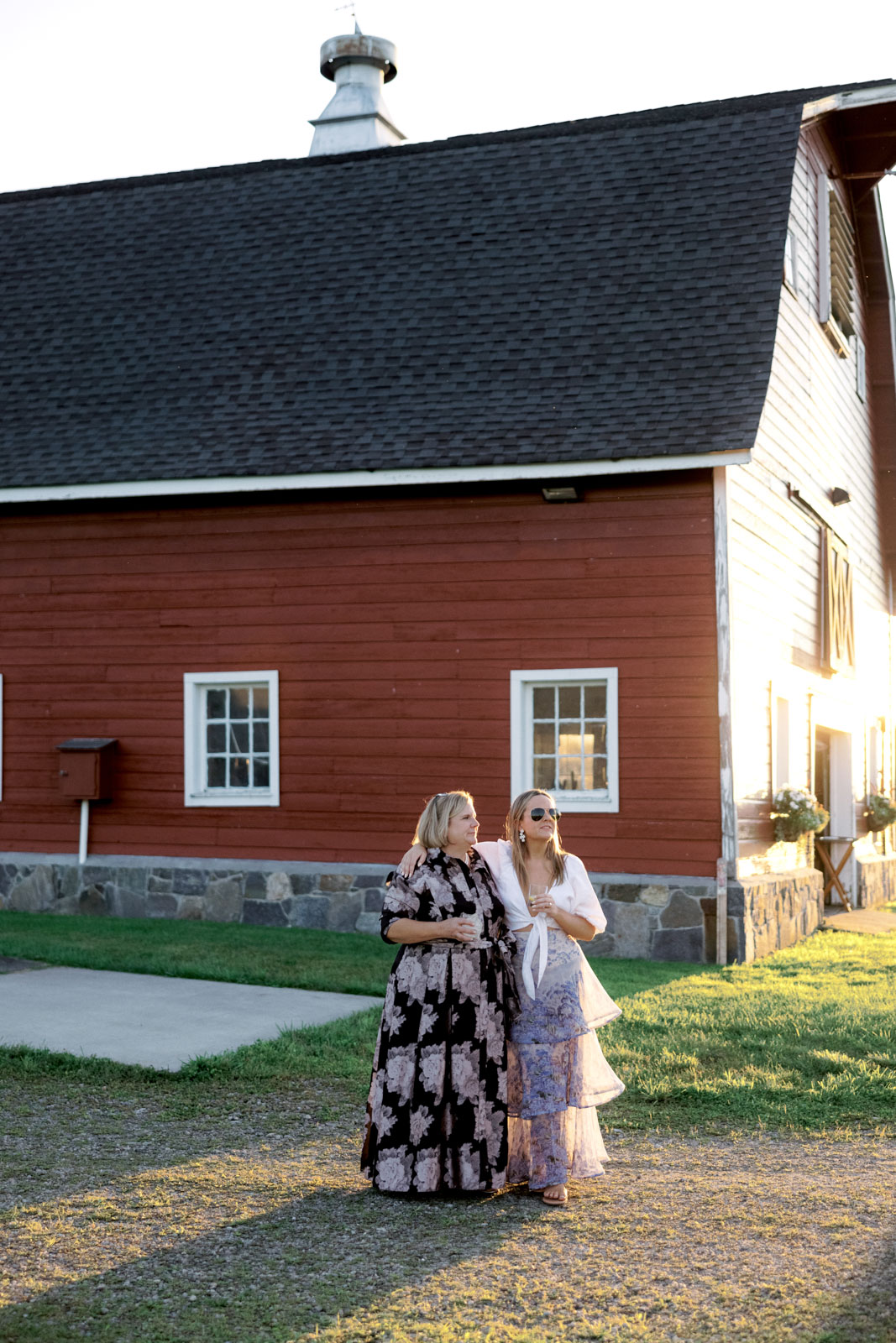 The bride's arm is on a lady's shoulder in front of a red barn house. Editorial rehearsal dinner image by Jenny Fu Studio.