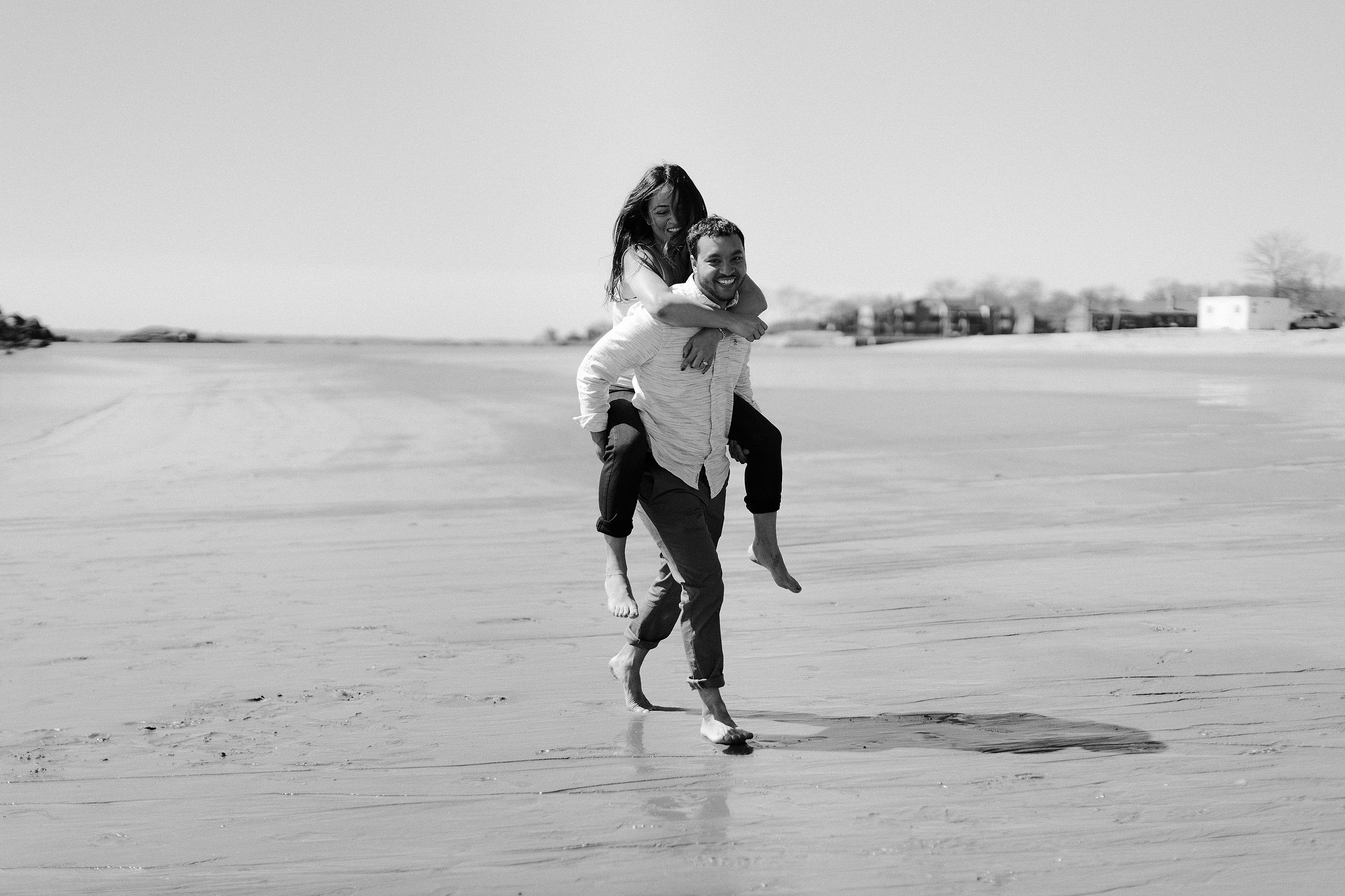 Black and white image of the man is carrying his fiancee on his back on the beach. Image by Jenny Fu Studio