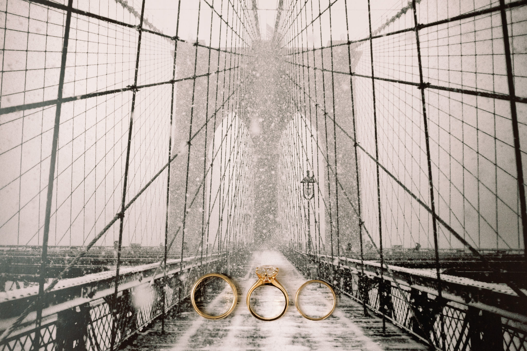 a set of wedding rings and an engagement band rest against the backdrop of the Brooklyn Bridge draped in a delicate blanket of snow. The timeless elegance of the rings is accentuated by the iconic bridge's architectural grace, creating a visual symphony that intertwines the enduring symbols of commitment with the ethereal beauty of a winter's day in New York.