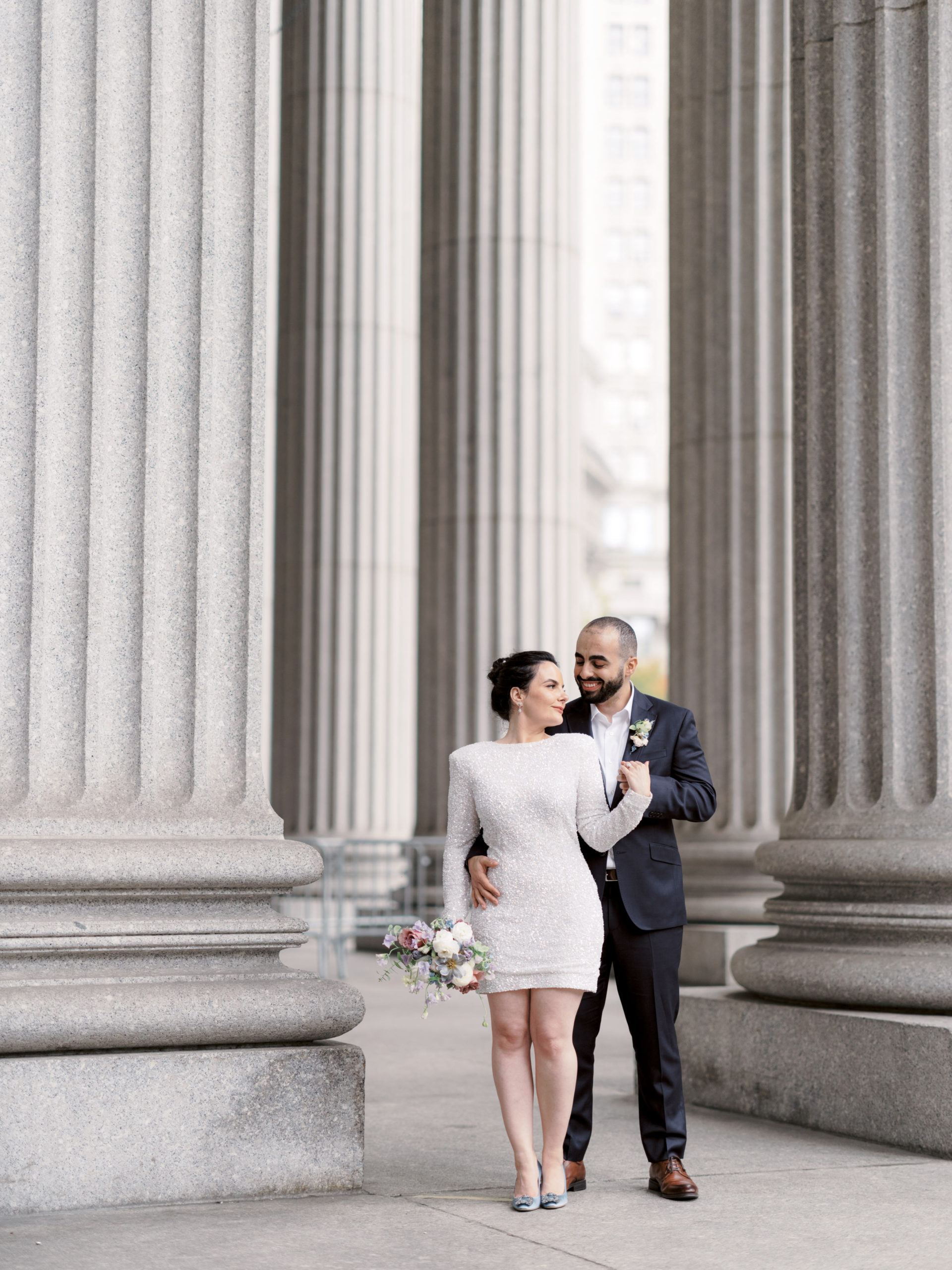The couple are standing in the middle of large columns in NYC City Hall Building. Editorial elopement Image by Jenny Fu Studio