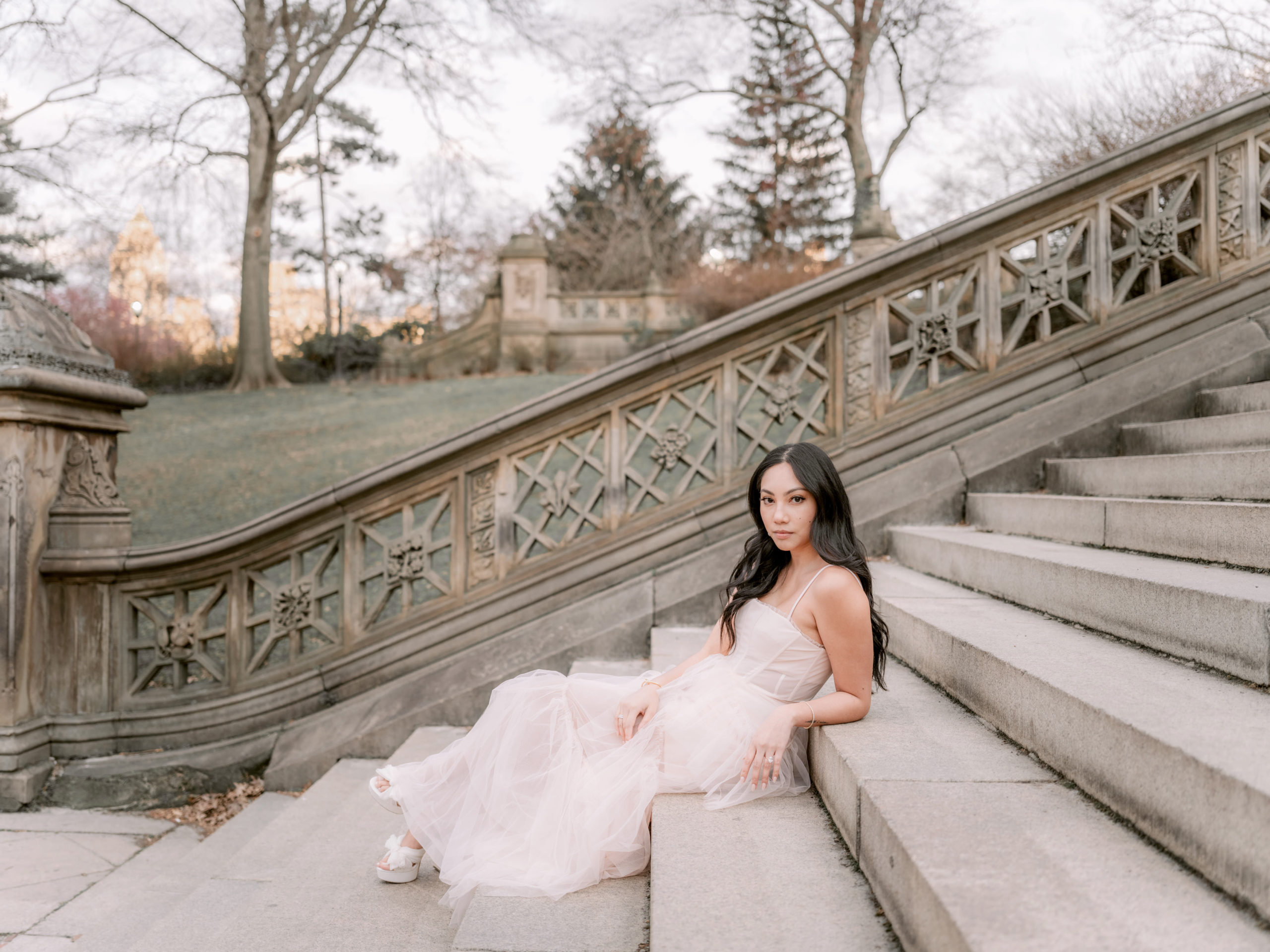 The engaged woman, wearing a beautiful engagement ring, is sitting on a staircase in Central Park. Editorial Photography by Jenny Fu Studio 