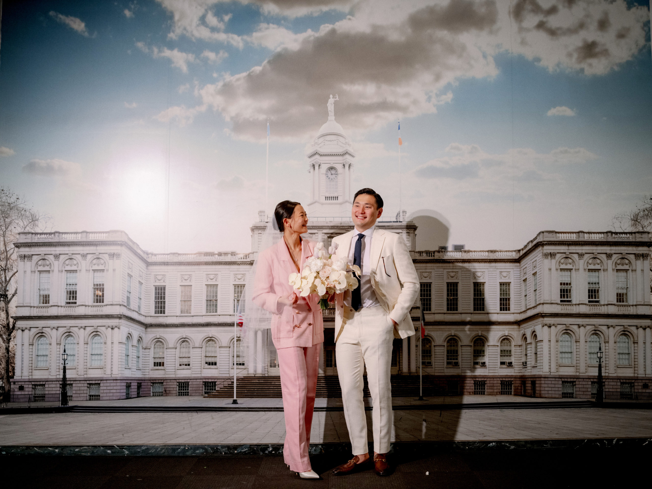 The newlyweds are laughing together, with a NYC City Hall photo in the background. Editorial elopement Image by Jenny Fu Studio