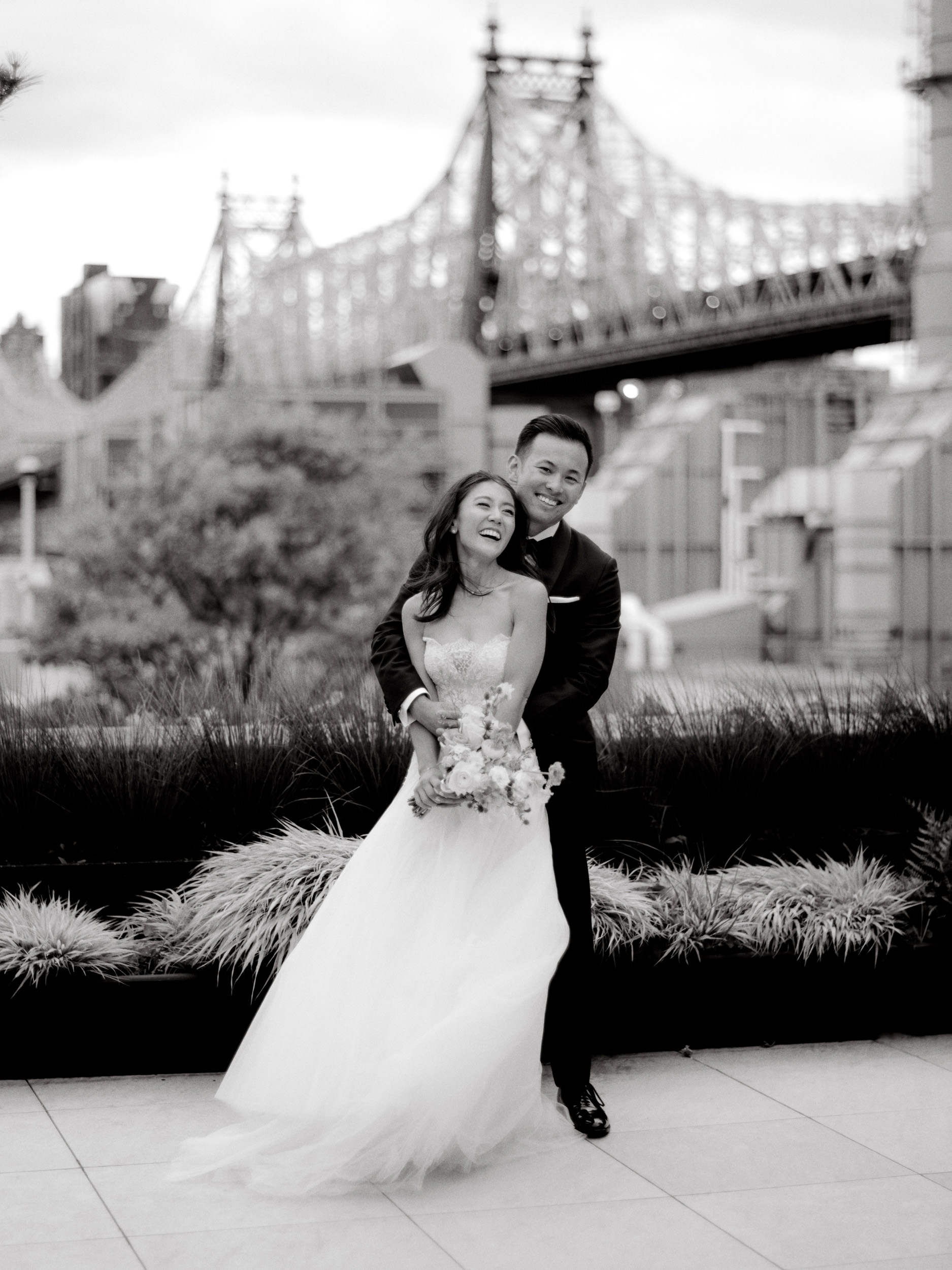 The bride and the groom are smiling happily with the brooklyn bridge on the background. Photojournalistic image by Jenny Fu Studio