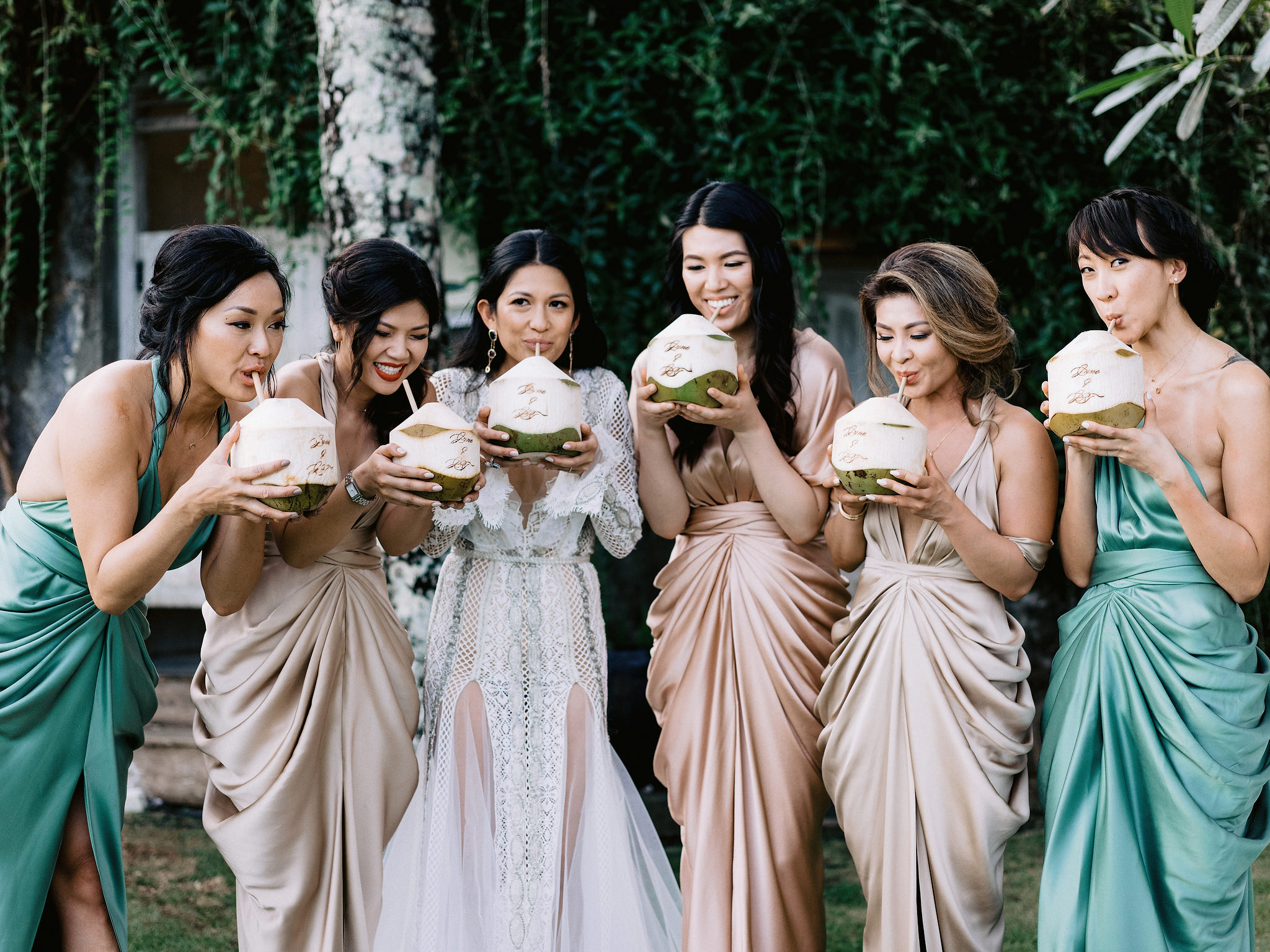 The bride and bridesmaids are drinking coconut water from the coconut fruit. Editorial destination wedding photo by Jenny Fu Studio
