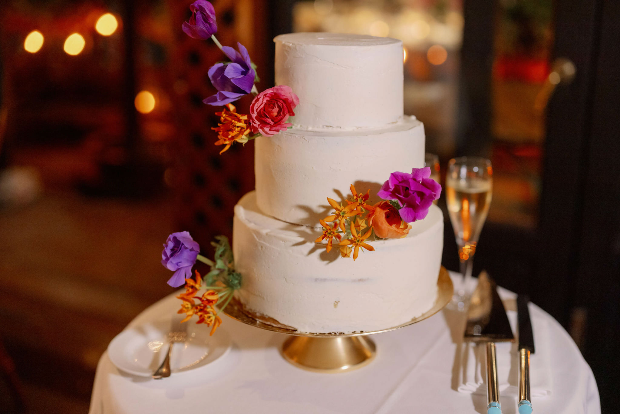 A three-layer white wedding cake with purple, orange and lavander-colored flowers. Editorial wedding image by Jenny Fu Studio