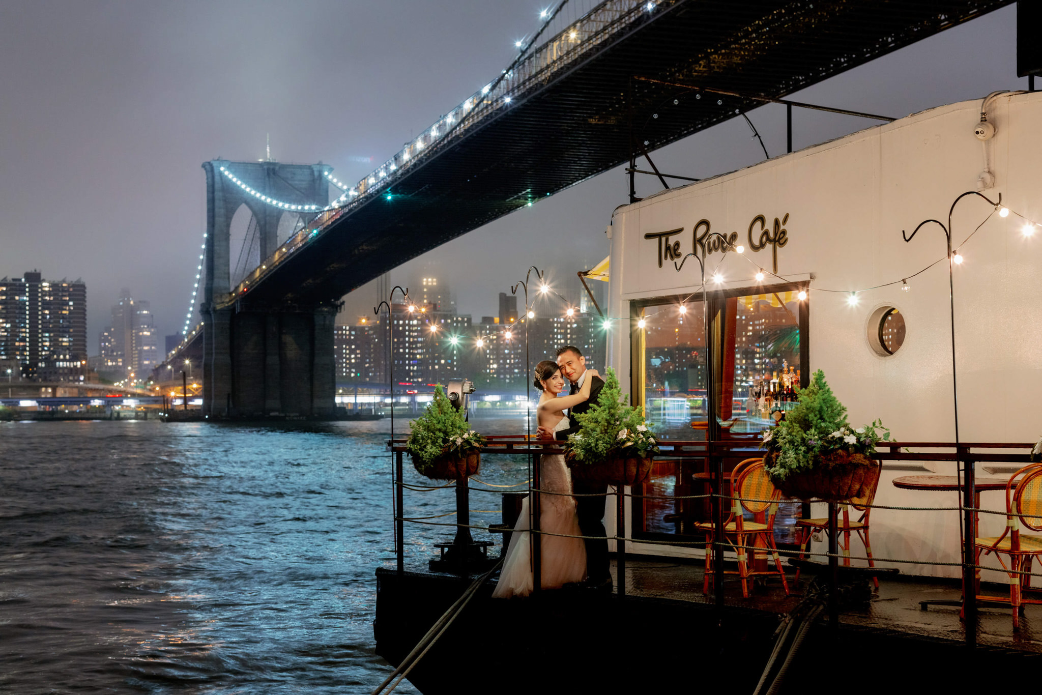 The newly-wed couple is standing in front of The River Cafe, with river and the Brooklyn Bridge in the background. Editorial wedding image by Jenny Fu Studio.