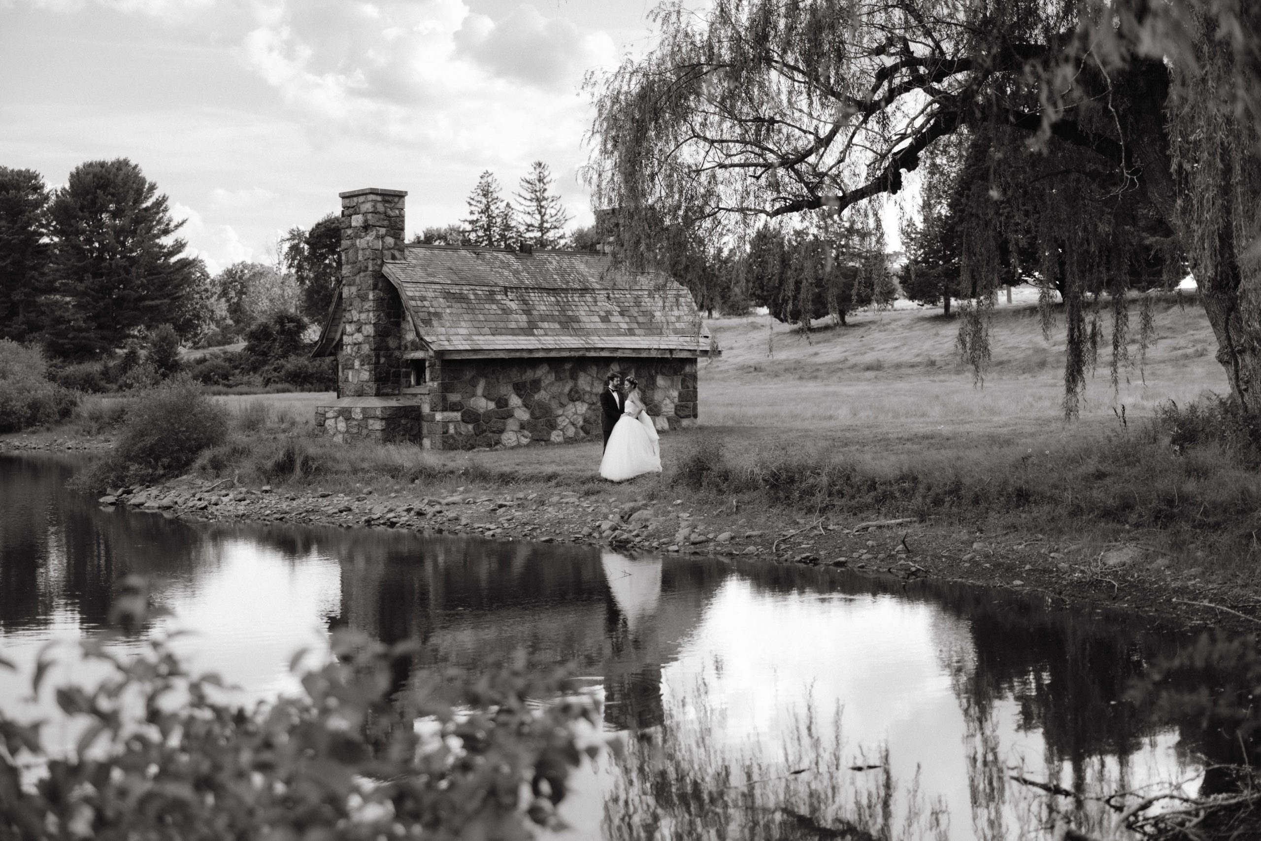 Black and white photo of the bride and groom in a lake with a cottage in the background. Image by Jenny Fu Studio