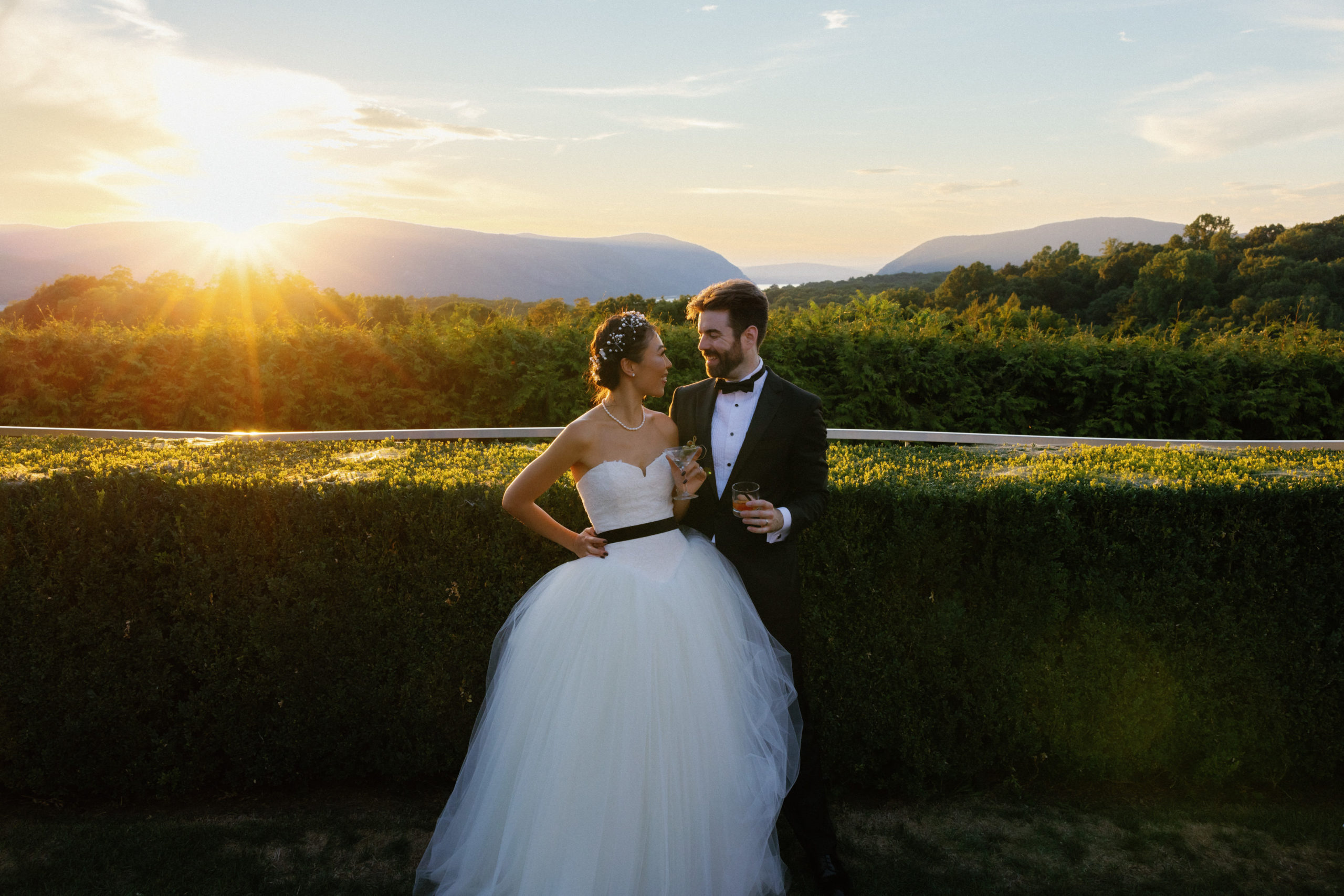 The bride and groom are smiling at each other while holding glasses of champagne. Sunset in the background. Image by Jenny Fu Studio