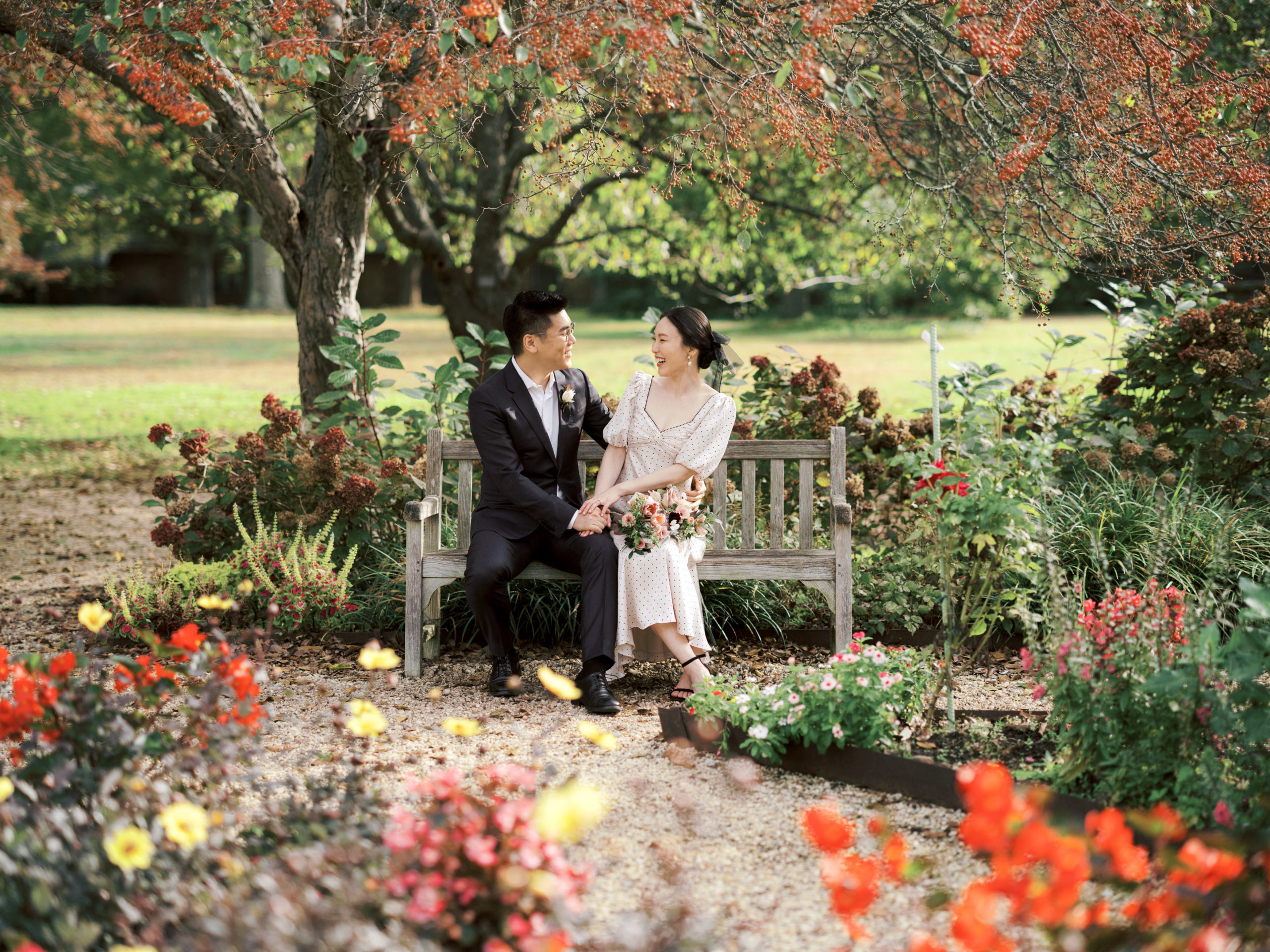 The engaged couple are having a good time, sitting at a bench, surrounded by beautiful flowers and tree. Engagement session photo by Jenny Fu Studio