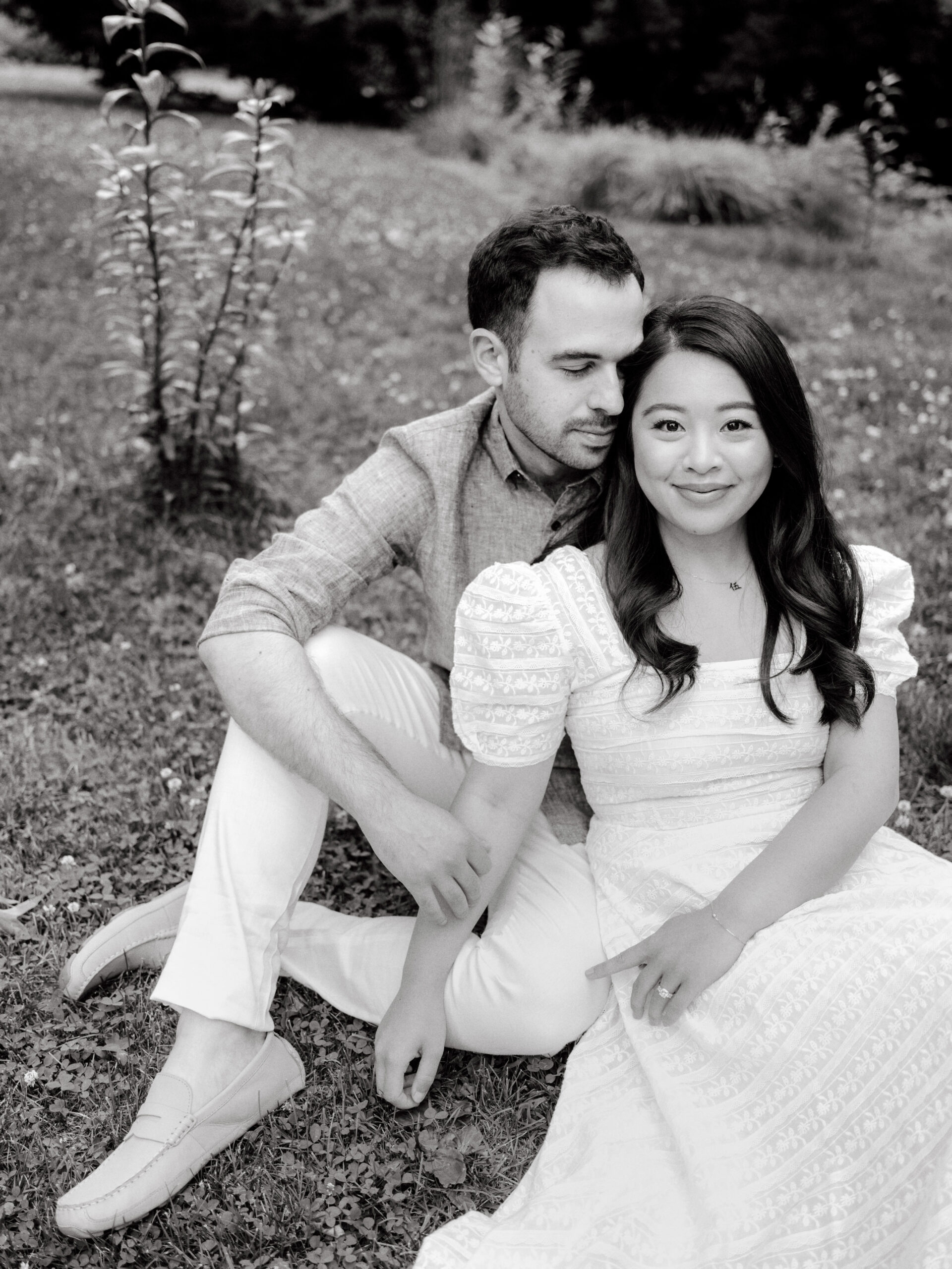 Black and white, editorial photo of the engaged couple sitting on the ground with nature in the background. Image by Jenny Fu Studio