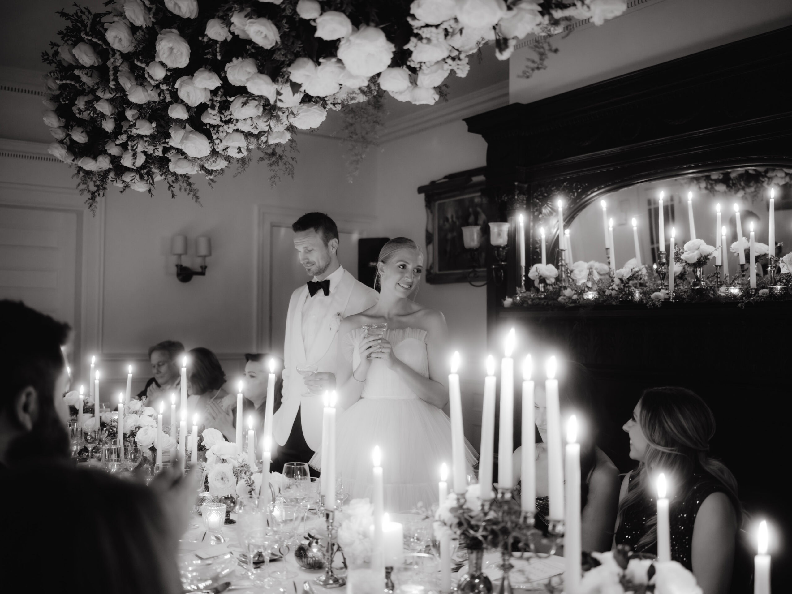 Black and white image of the bride and groom standing beside the dining table while the guests are eating. Image by Jenny Fu Studio