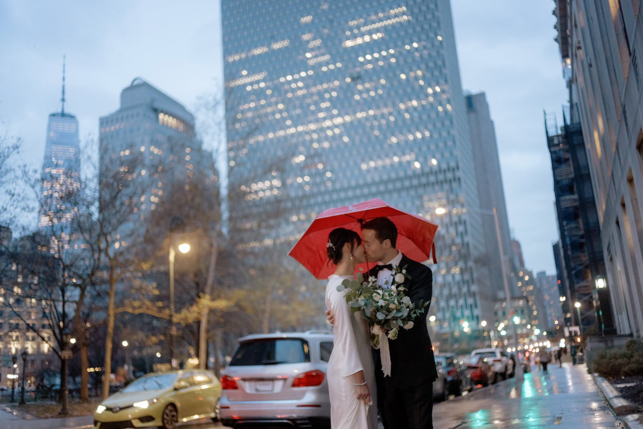 a couple shares a tender kiss under the shelter of a red umbrella, surrounded by the soft glow of city lights during the bewitching blue hour. Following their New York City Hall elopement, the rain-kissed streets add a poetic touch to the scene, weaving an intimate tapestry of love against the urban backdrop