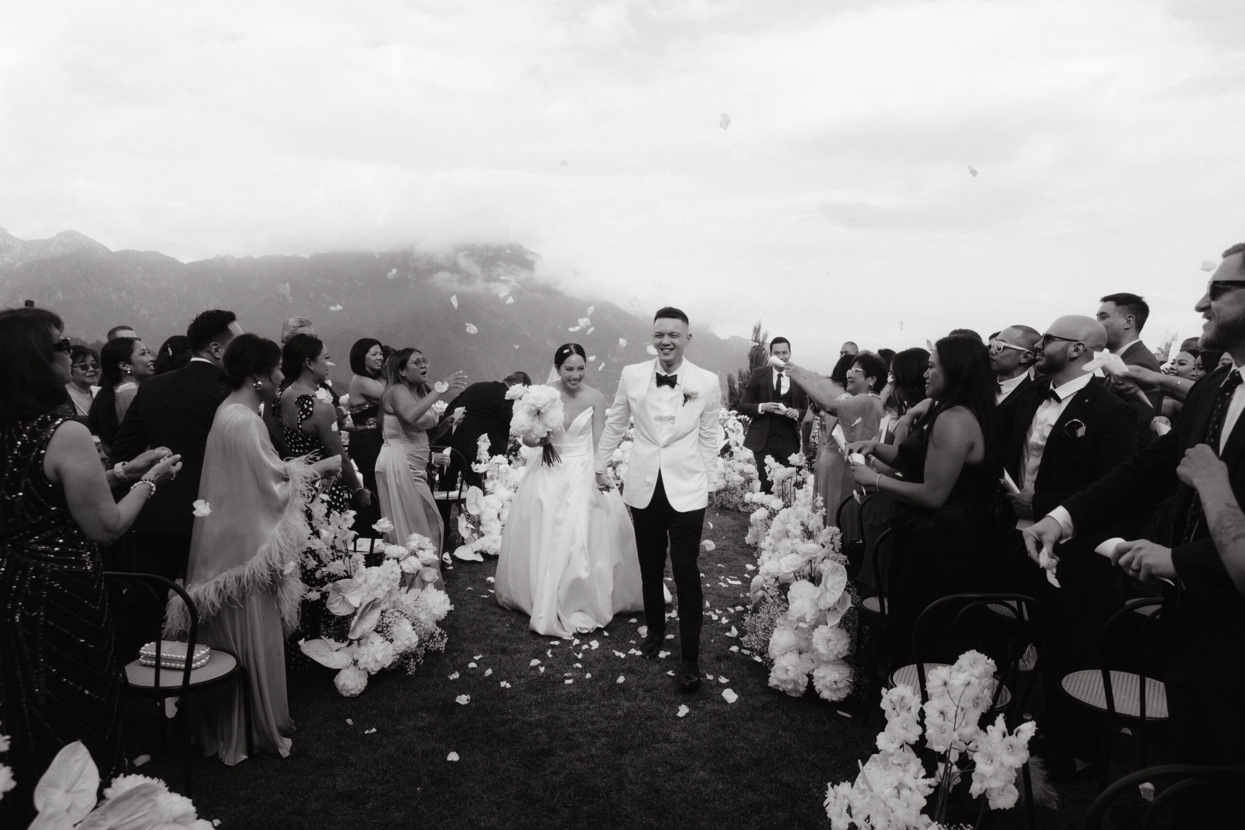 In a timeless black and white photo, the bride and groom joyfully exit their ceremony, bathed in white rose petals. Cheers from guests surround them against the breathtaking backdrop of the Amalfi Coast, with clouds hovering over majestic mountains. A moment of pure happiness captured in a picturesque celebration.