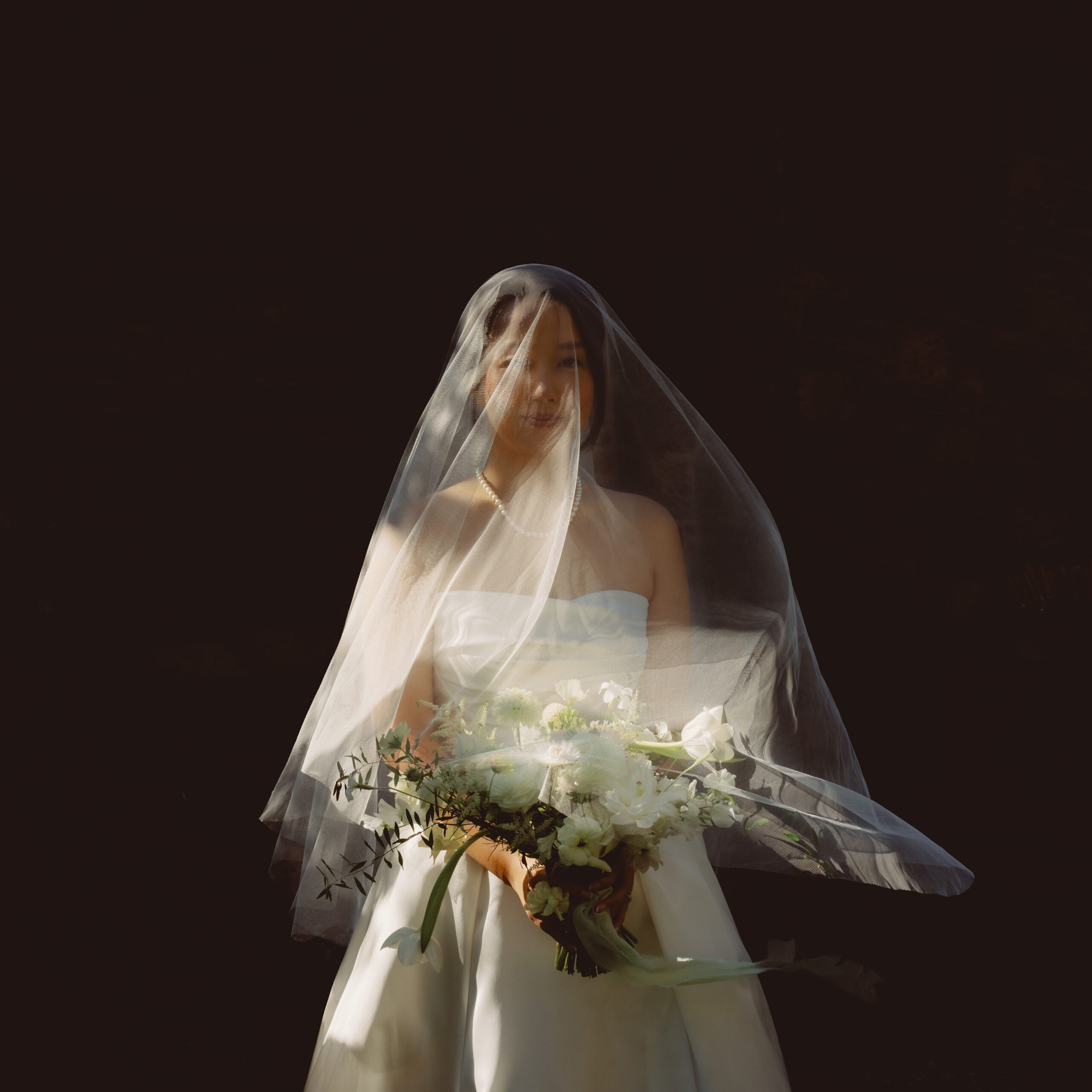 Captivating fine art photograph of a bride, adorned with a veil and holding flowers, bathed in soft sunlight. The scene, resembling a masterpiece, was artistically captured by Jenny Fu at Blue Hill at Stone Barns in New York, evoking the timeless elegance of a painting brought to life.