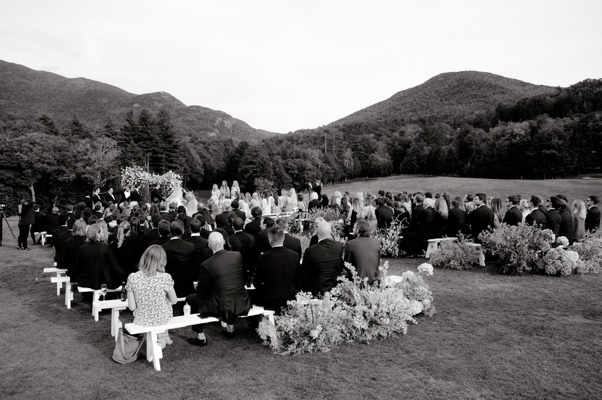 Scenic wedding ceremony atop a hill in the Adirondack Mountains. The couple exchanges vows surrounded by nature's grandeur, with panoramic views of majestic mountains creating a stunning backdrop for their special day.