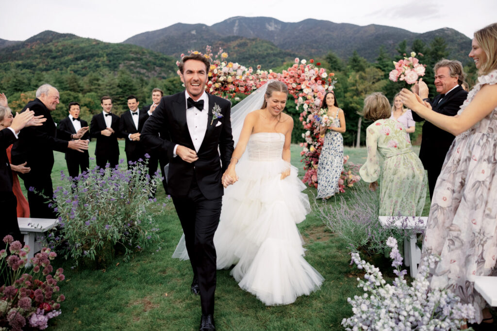 A timeless image of pure happiness as the bride and groom exit their wedding ceremony in the Adirondack Mountains. The couple wears beaming smiles while guests cheer in celebration, surrounded by flowers, joy, and love. Captured beautifully by photographer Jenny Fu, this moment encapsulates the essence of a joyous and celebratory wedding day.