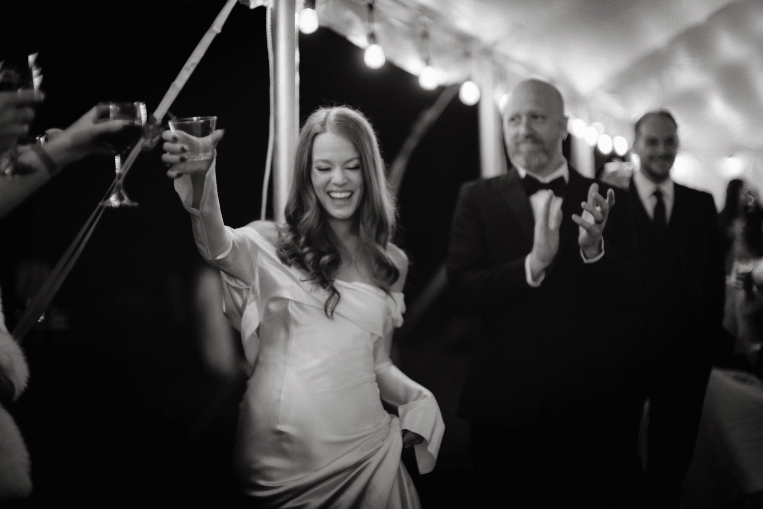 Black and white photo of the bride happily doing a toast with guests while the groom is clapping his hand in the background. Image by Jenny Fu Studio