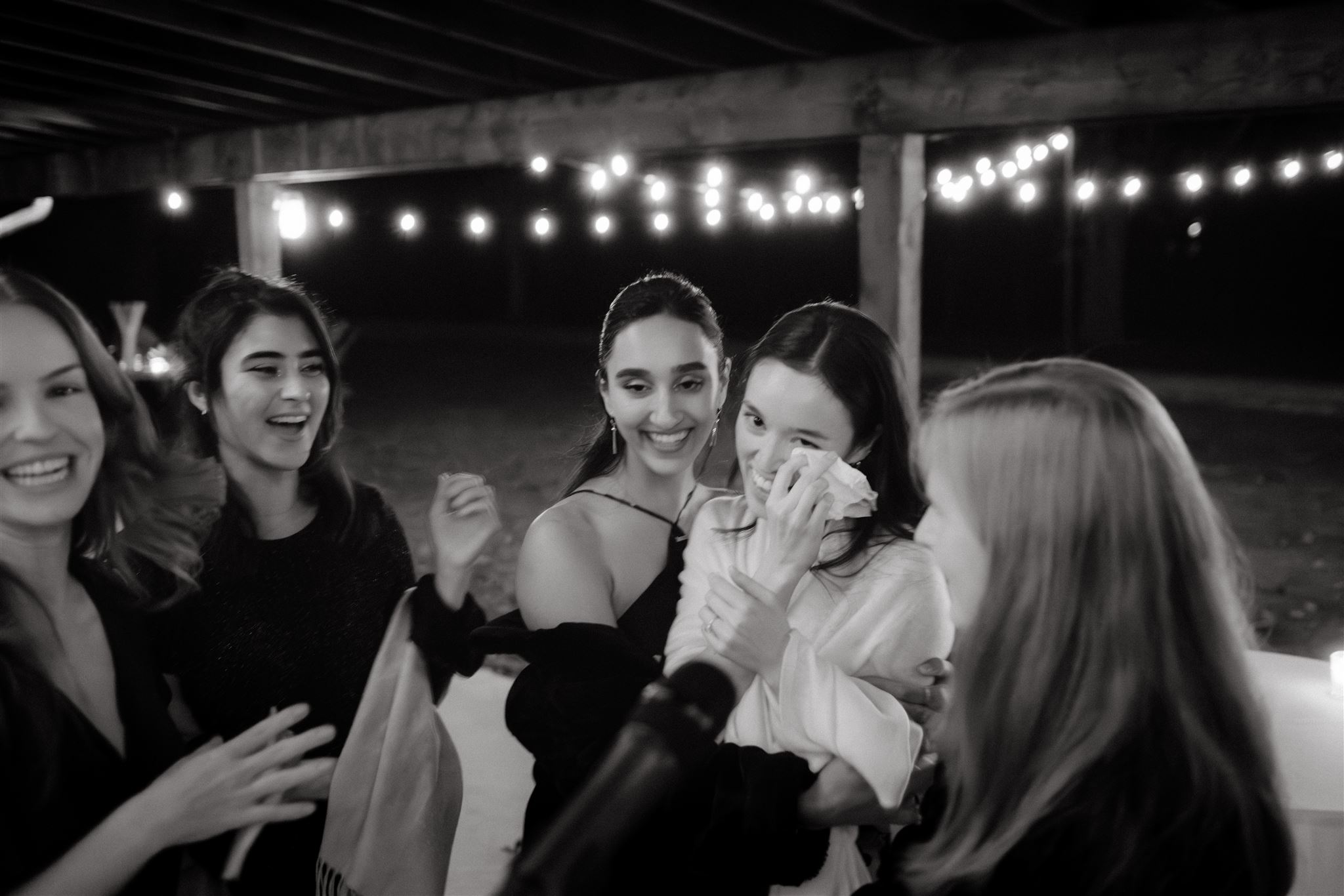 Black and white candid image of the bride wiping her tears while smiling, surrounded by her friends. Image by Jenny Fu Studio