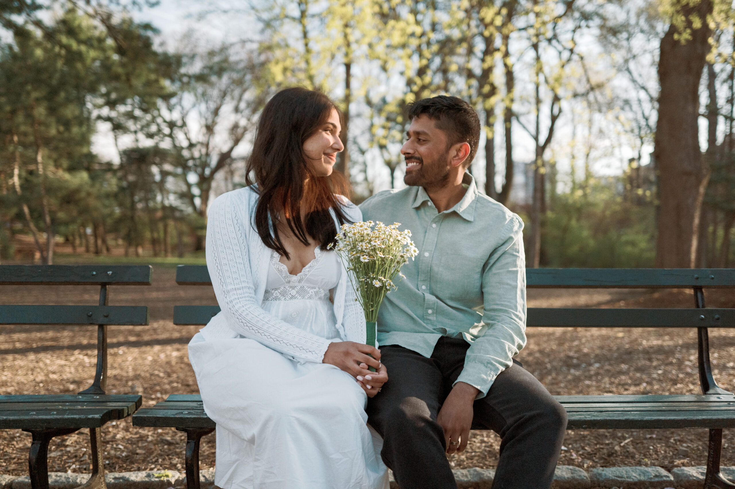 The engaged couple are looking into each other's eyes while sitting on a bench in a park outdoors. Engagement sessions image by Jenny Fu Studio