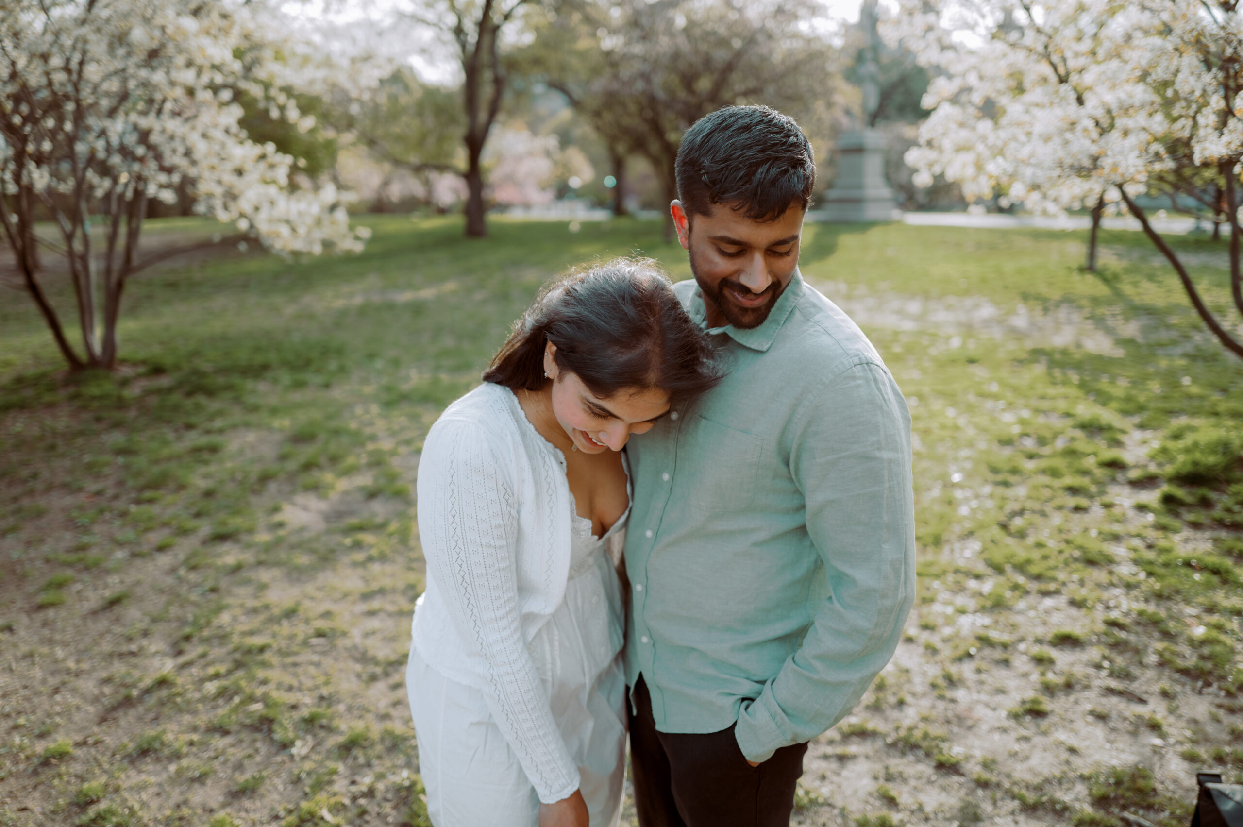 The engaged couple are happily looking downwards in a park during spring time. Engagement sessions image by Jenny Fu Studio