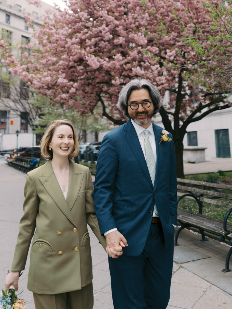 The newlyweds are walking hand in hand in NYC after their spring time wedding with cherry blossoms in the background. Image by Jenny Fu Studio