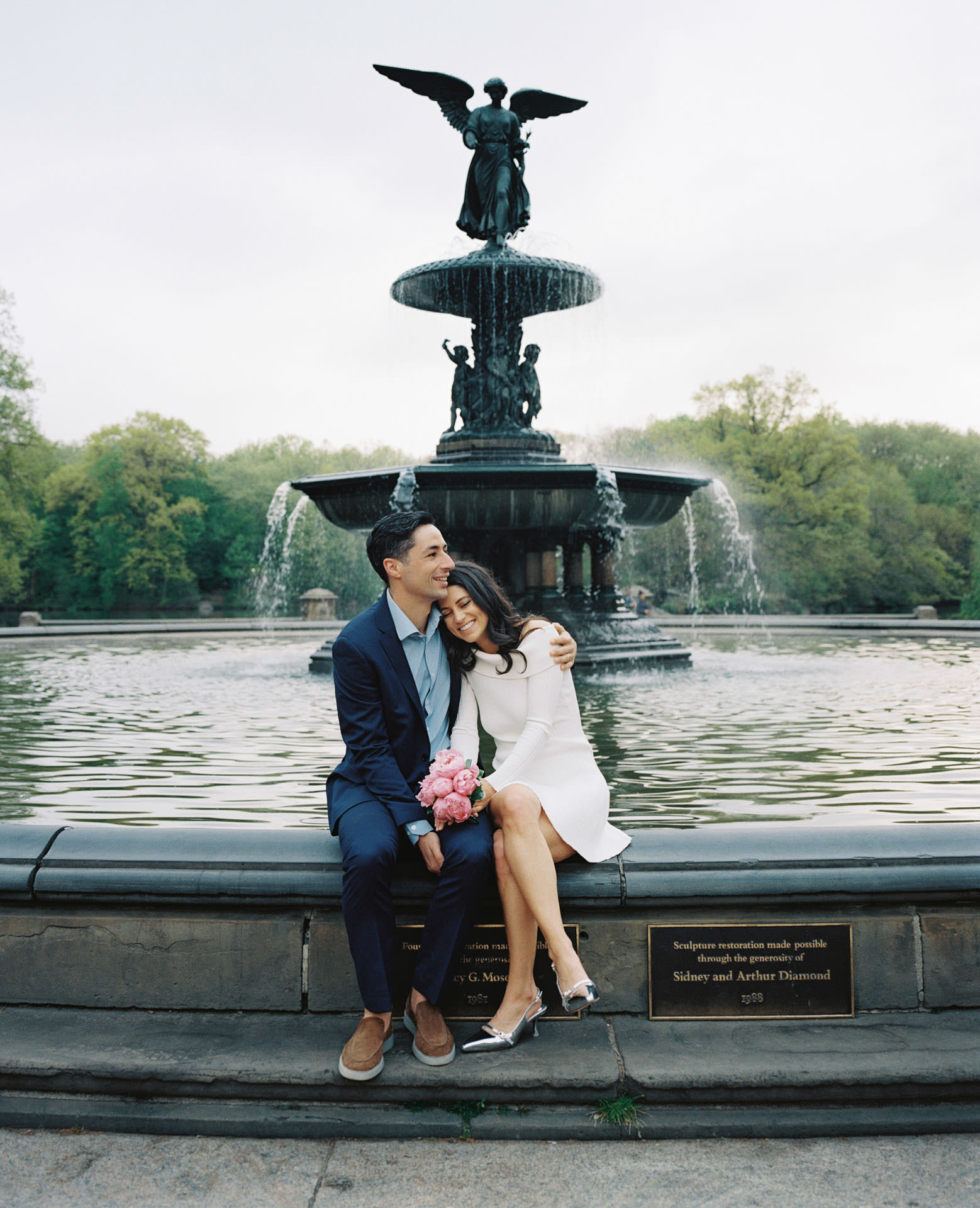 An engaged couple posing lovingly in front of the Bethesda Fountain, with water cascading in the background.

